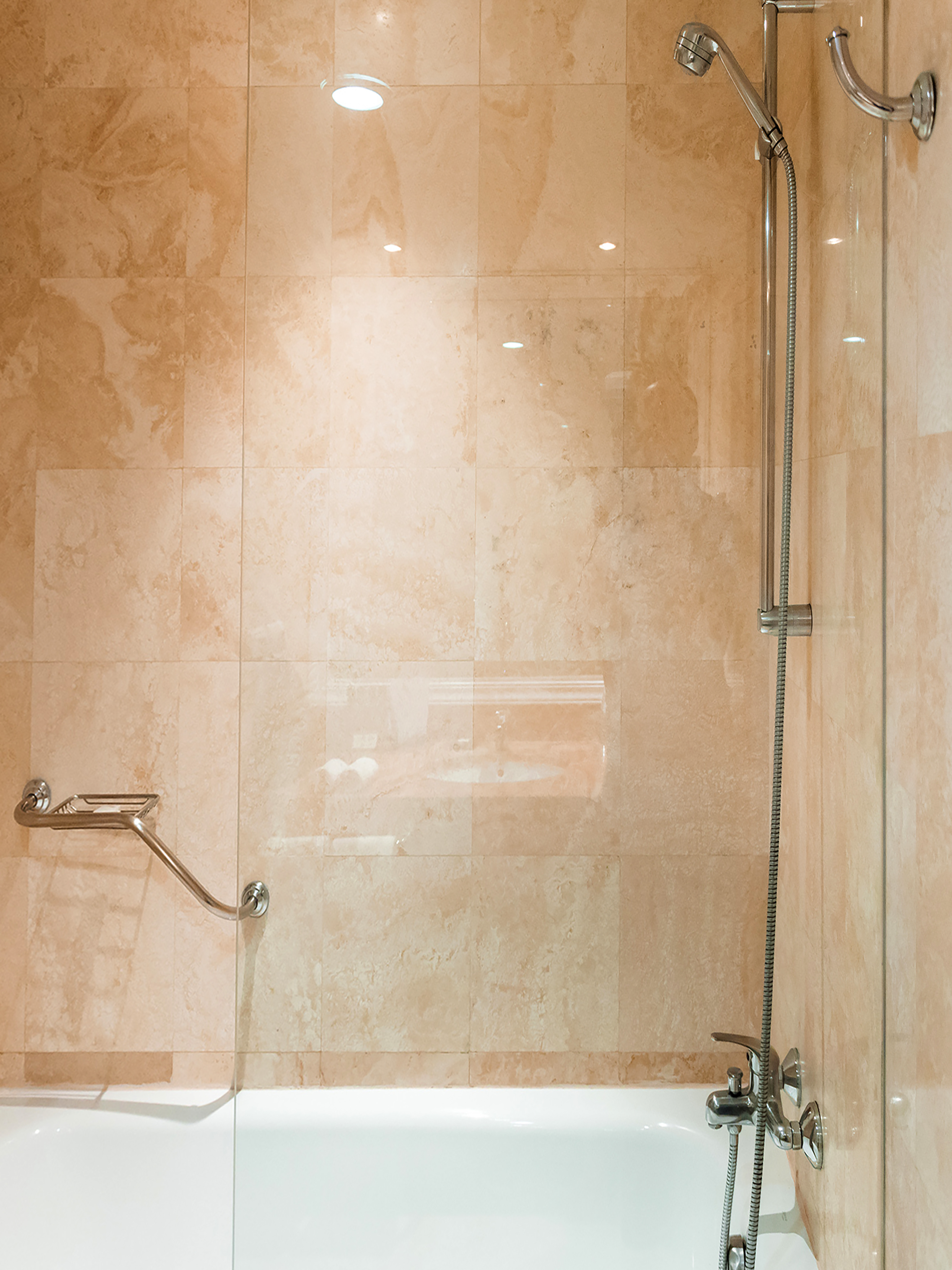 Bathtub and shower with beige marble tiles, handheld shower, and safety grab bar.