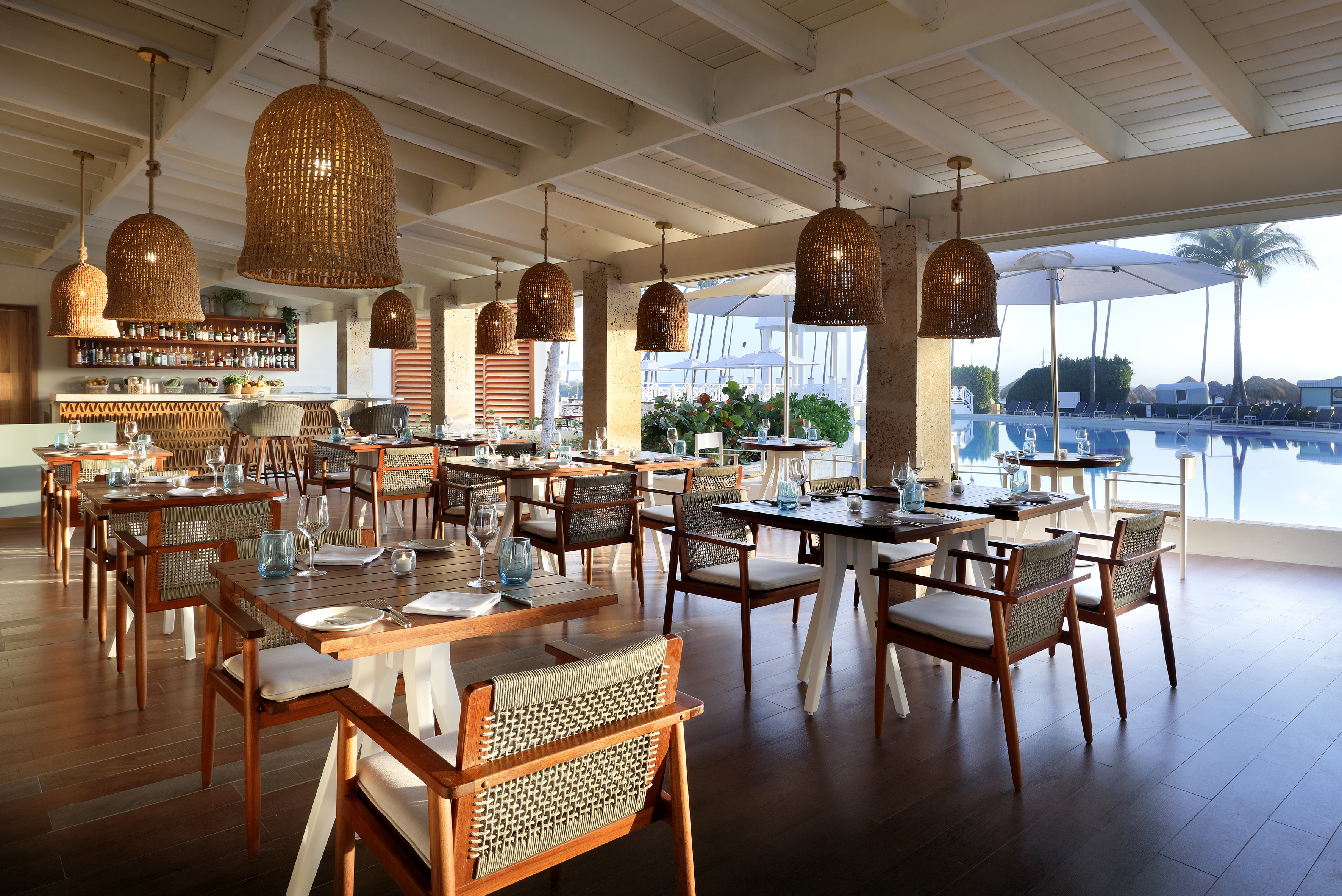 Tropical resort restaurant with wooden tables, wicker lights, and poolside view.