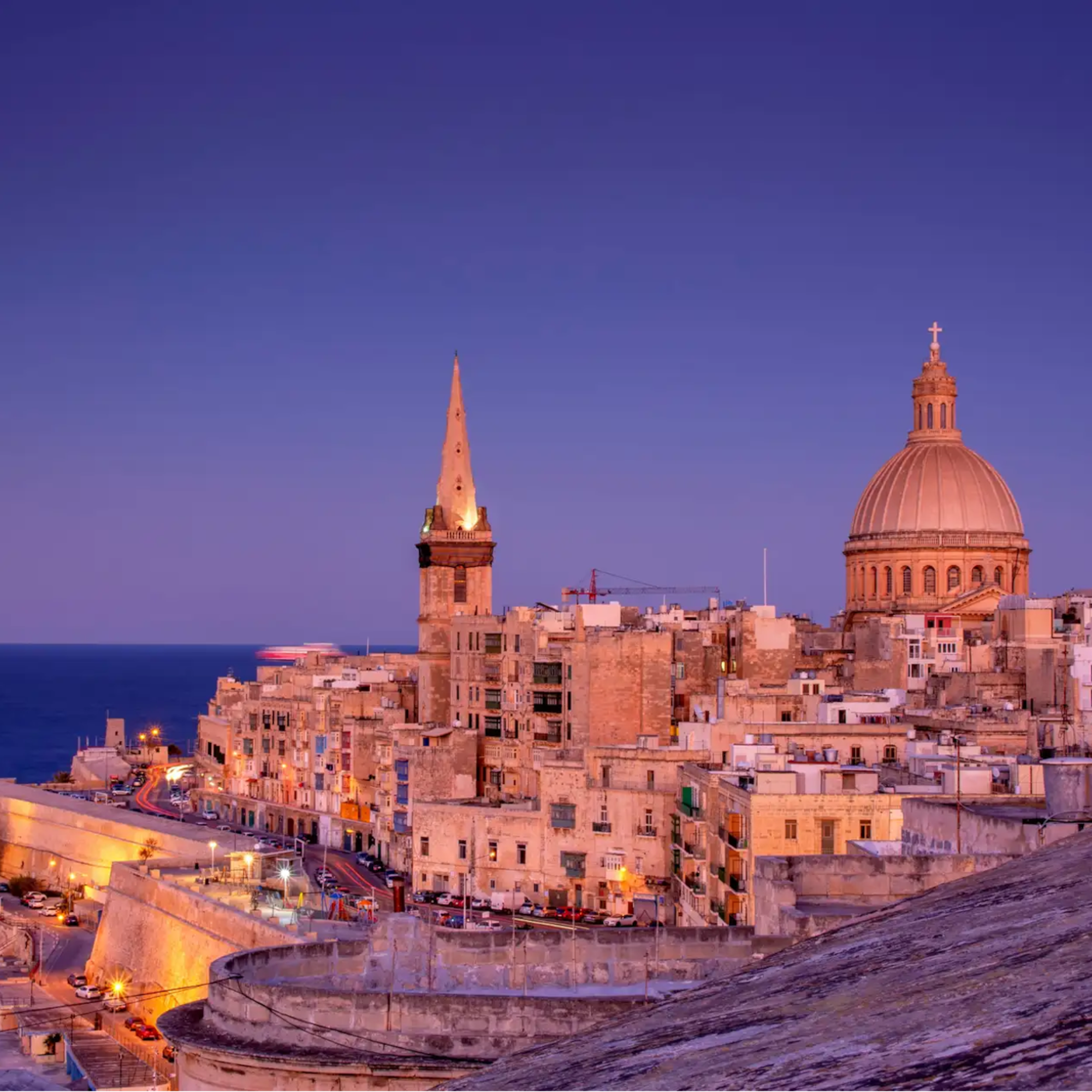 Historic Valletta skyline at twilight with illuminated dome, steeple, and coastline.