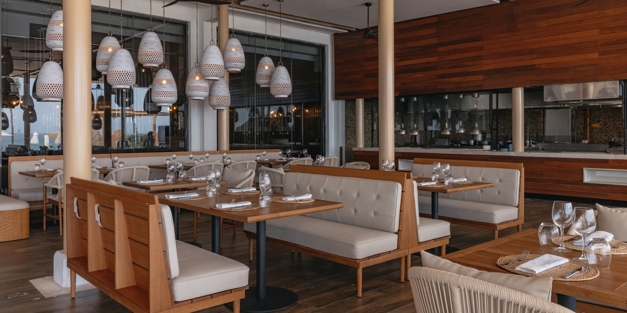 Modern restaurant with wood tables, beige booths, white pendant lights, open kitchen.