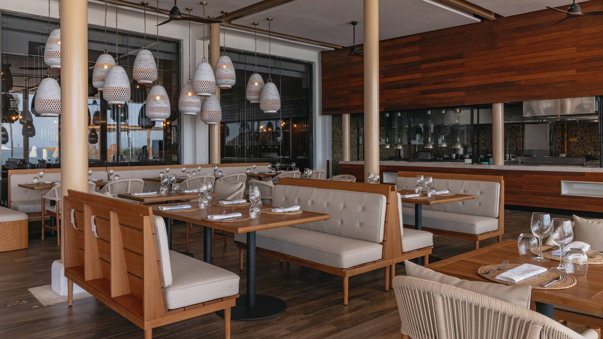 Modern restaurant with wood tables, beige booths, white pendant lights, open kitchen.