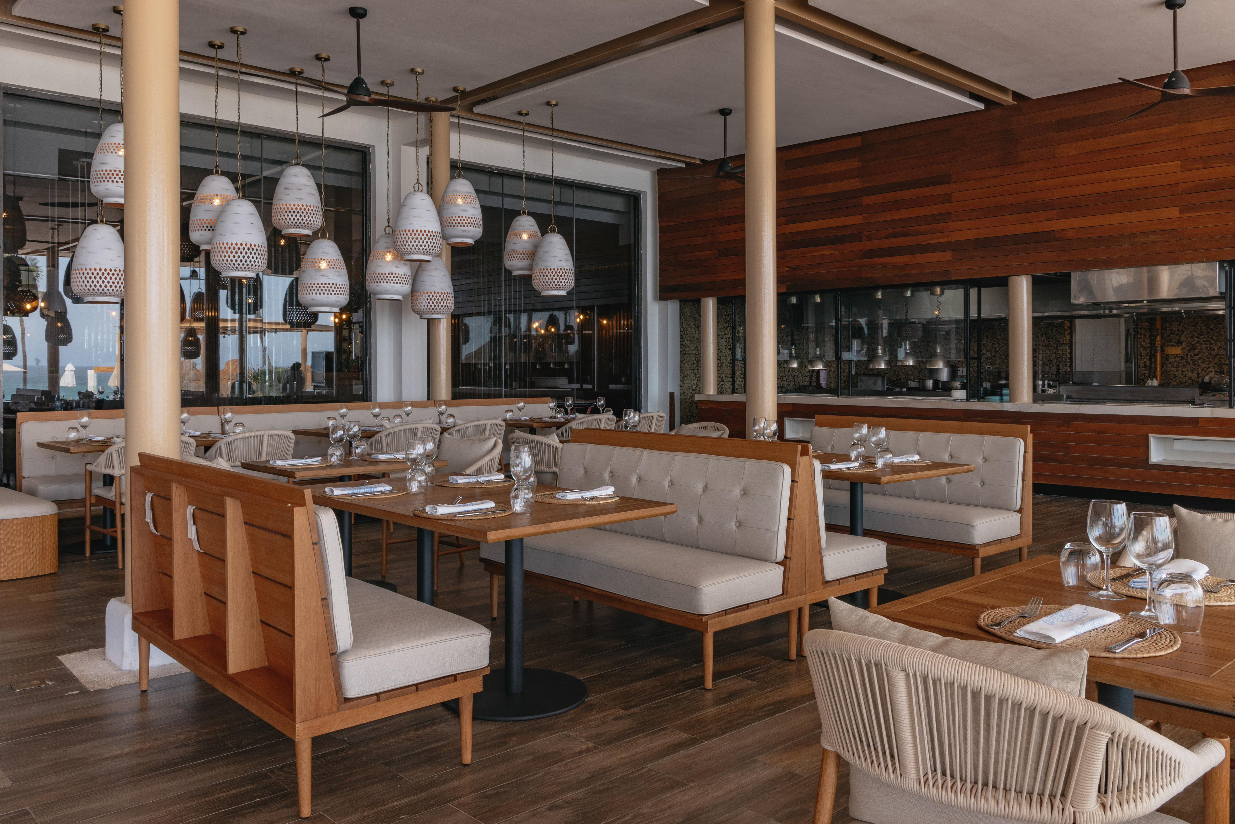 Modern restaurant with wood tables, beige booths, white pendant lights, open kitchen.