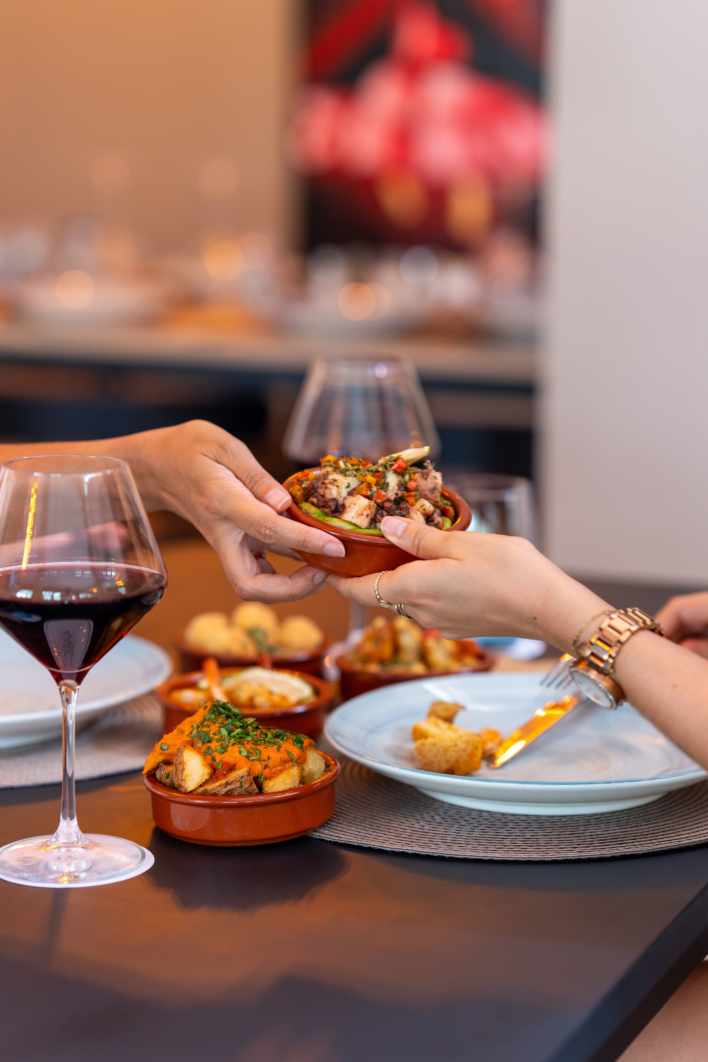 Hands sharing octopus tapas at a restaurant table with red wine and patatas bravas.