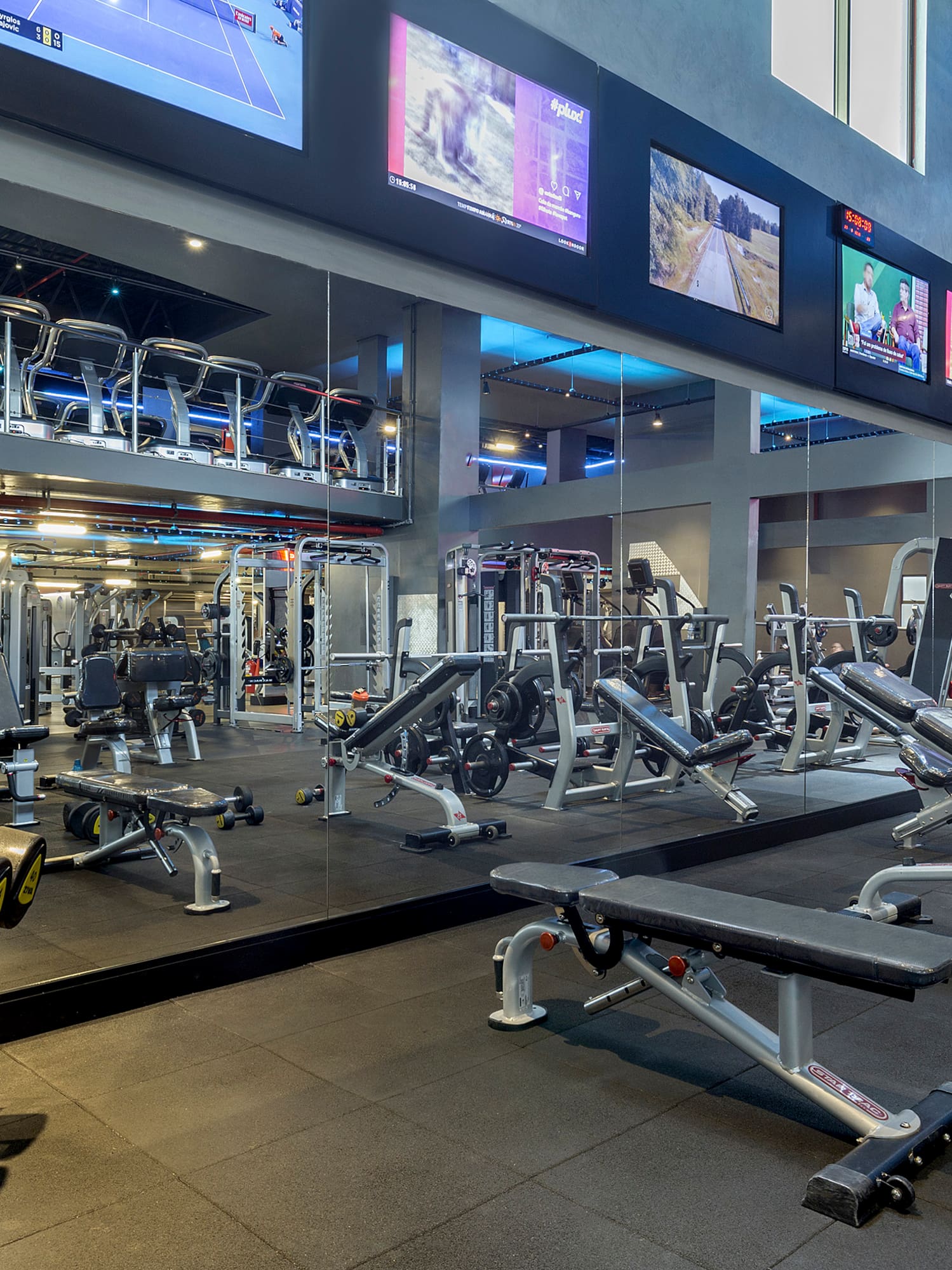 Modern fitness center with rows of dumbbells, weight machines, and large screens.