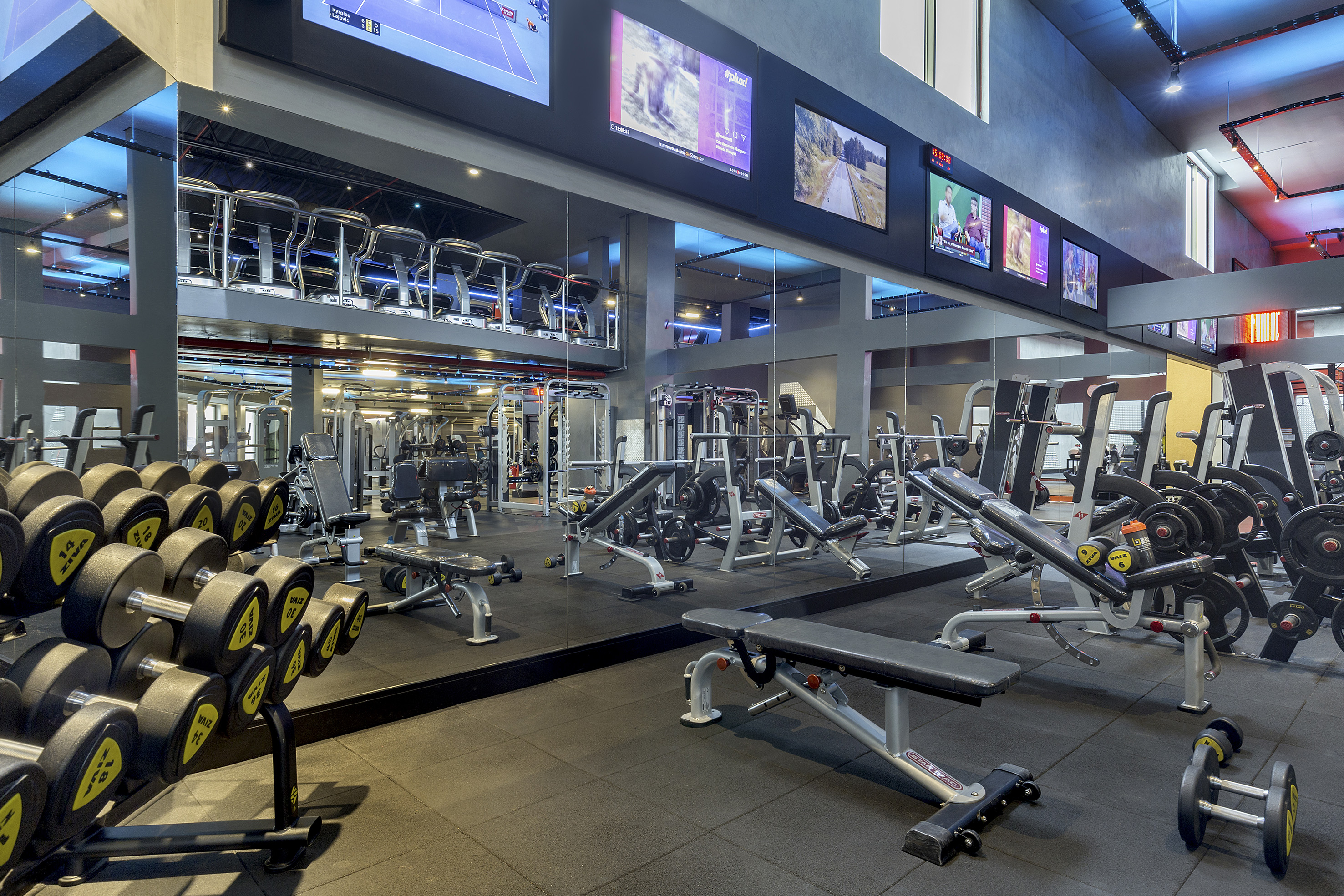 Modern fitness center with rows of dumbbells, weight machines, and large screens.