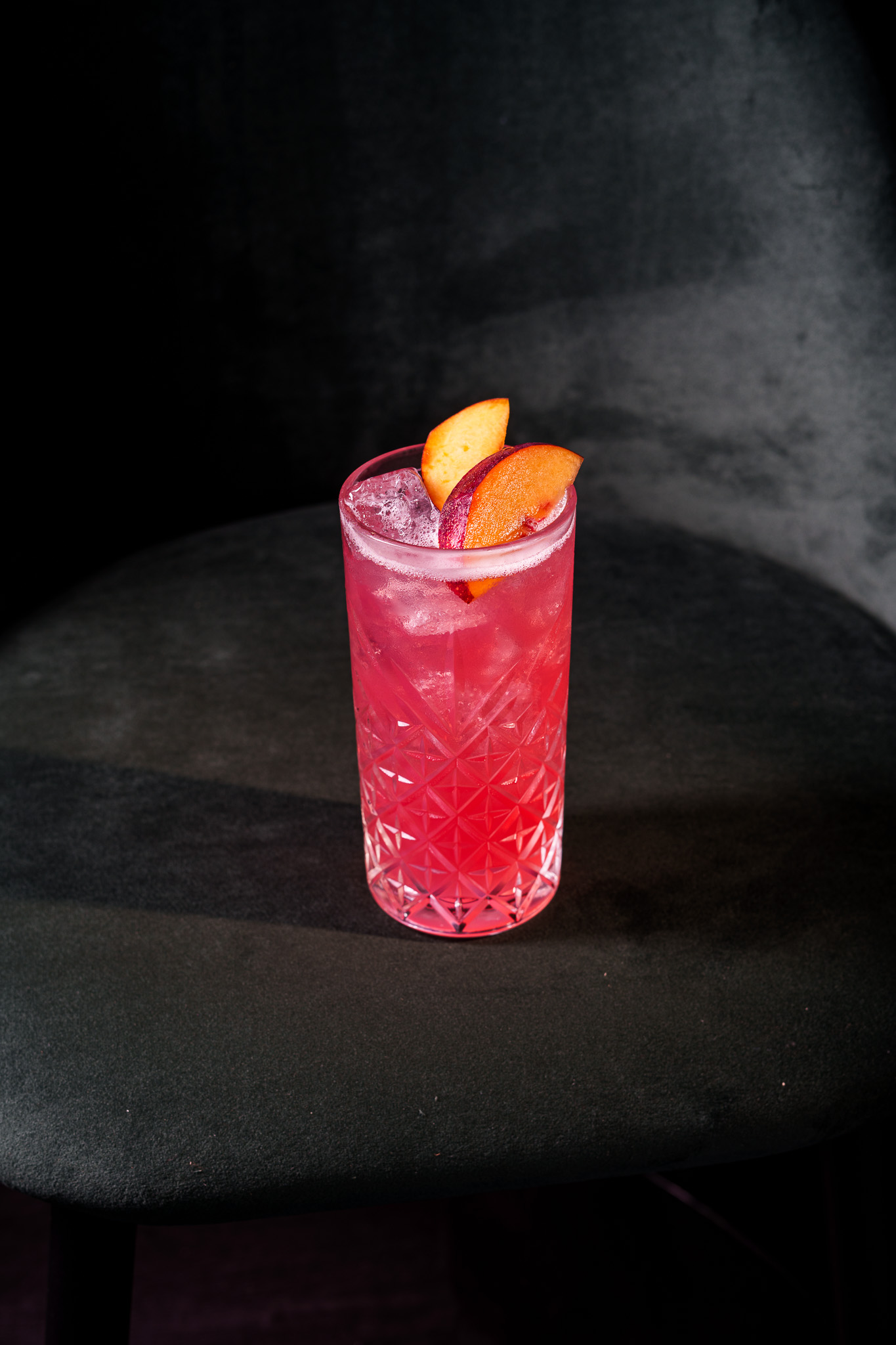 Iced pink cocktail in textured glass, garnished with peach and plum slices.