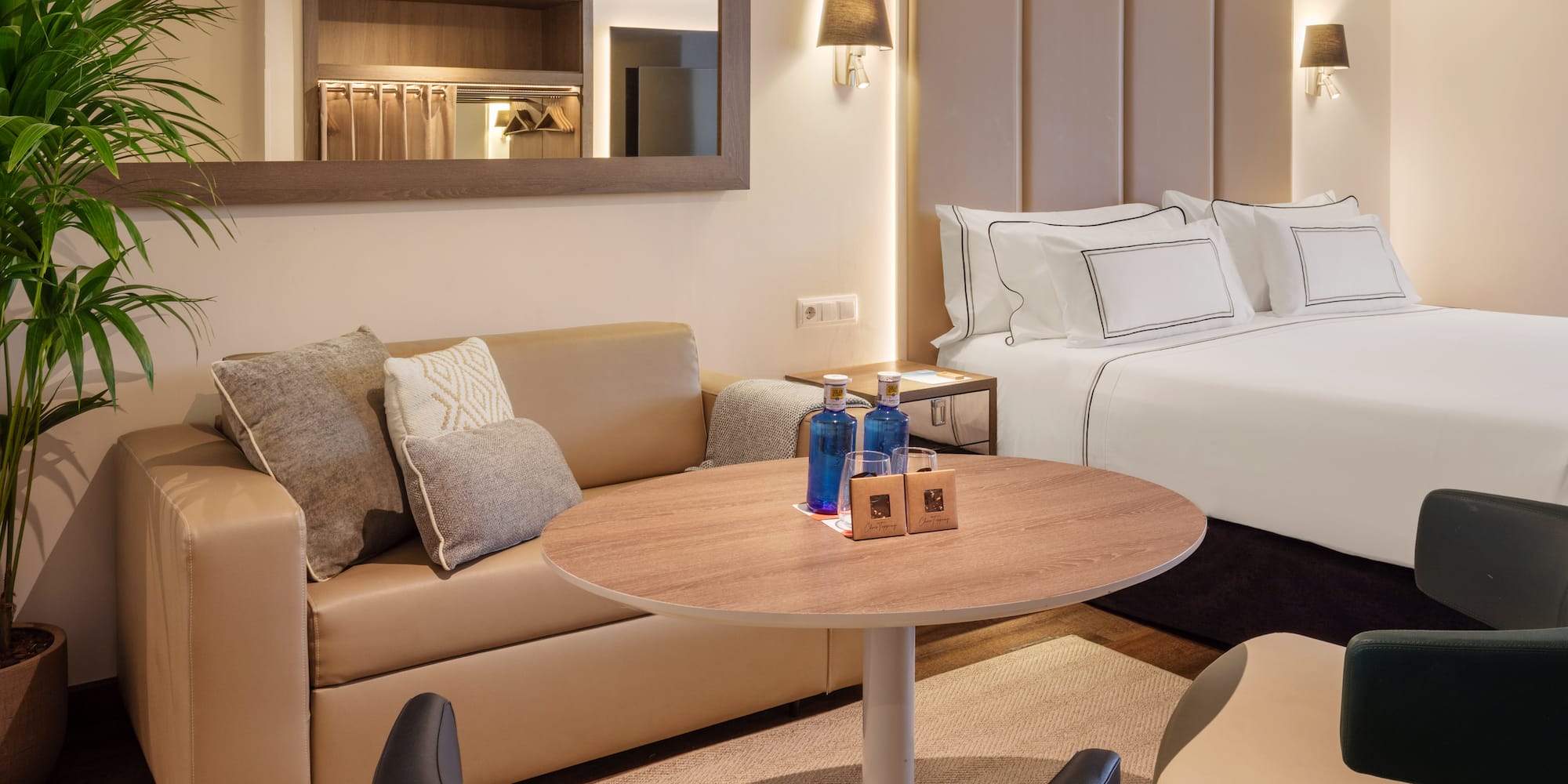 Modern hotel suite with large bed, beige sofa, round table, and bottled water.