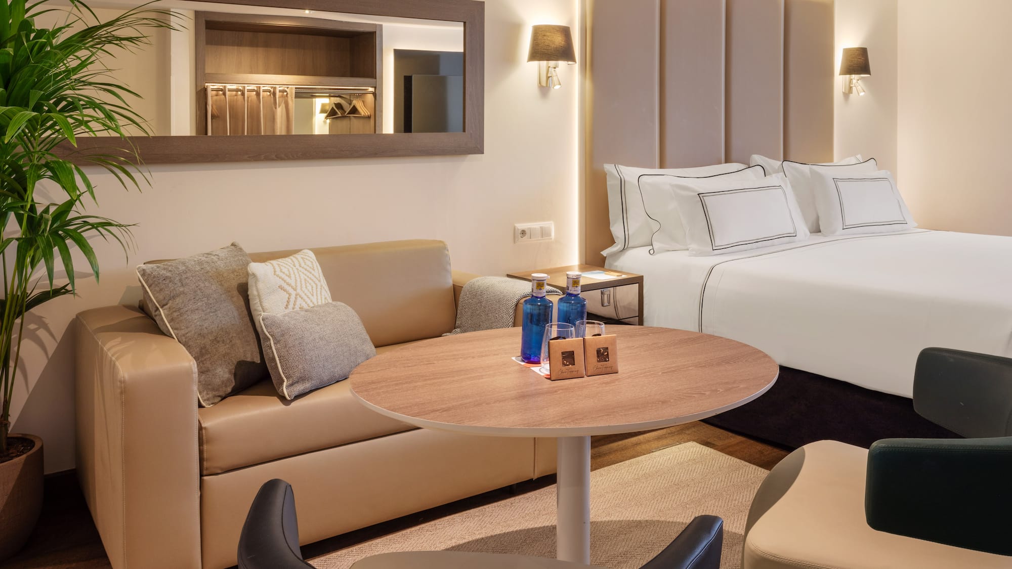 Modern hotel suite with large bed, beige sofa, round table, and bottled water.