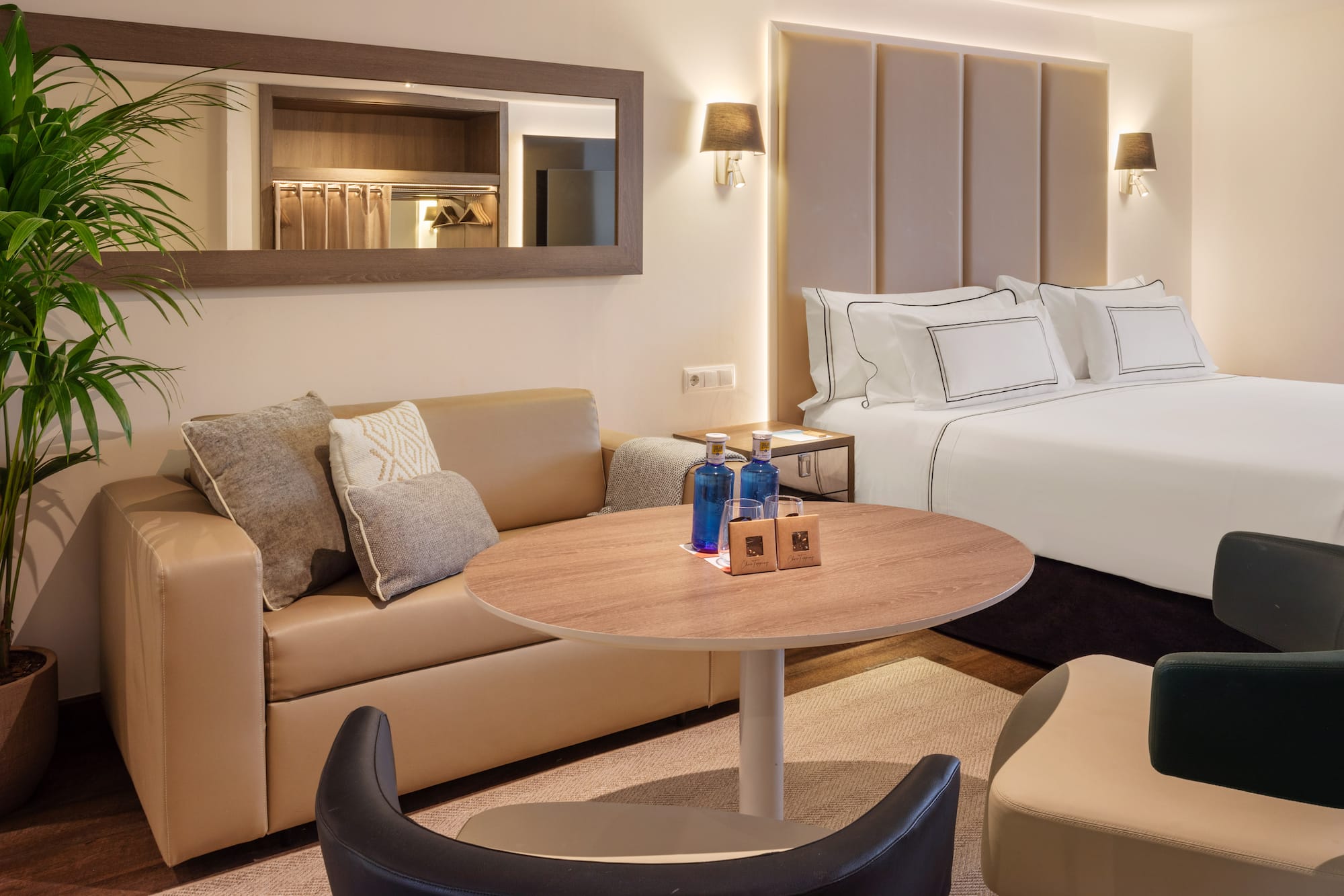 Modern hotel suite with large bed, beige sofa, round table, and bottled water.