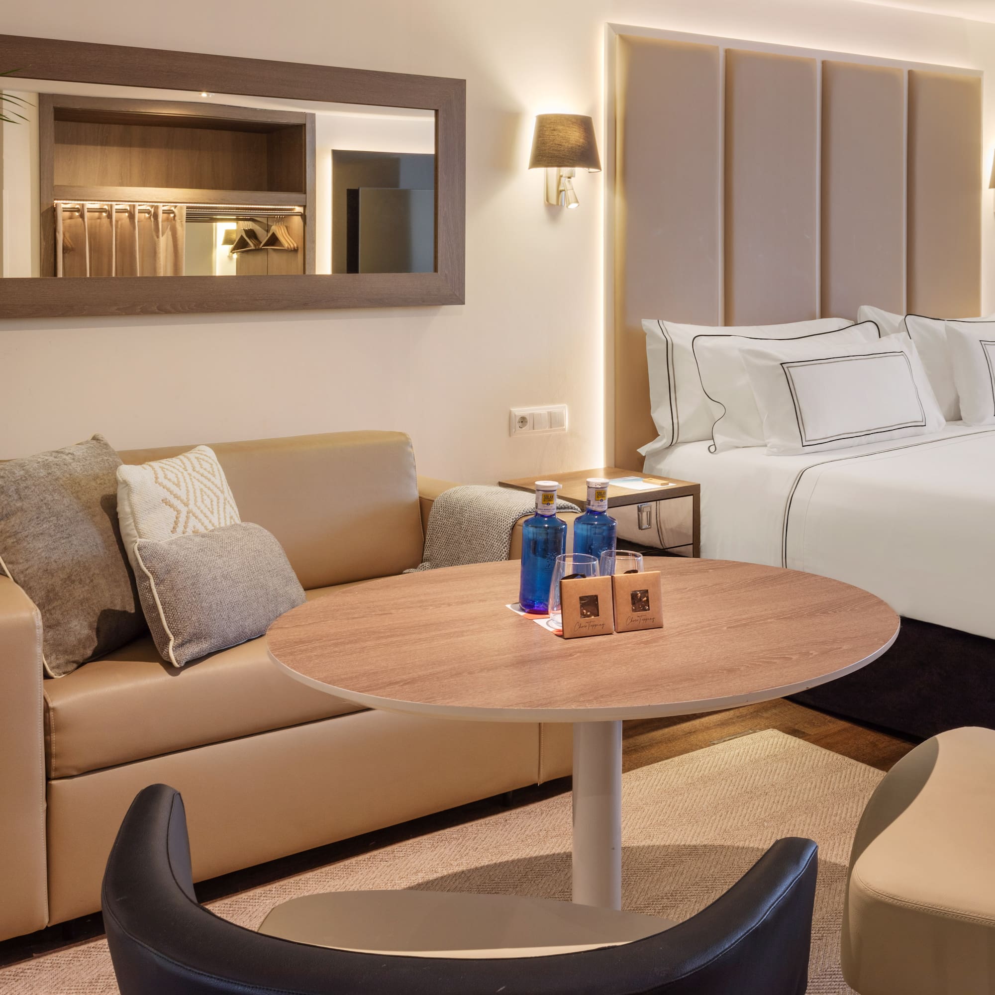 Modern hotel suite with large bed, beige sofa, round table, and bottled water.