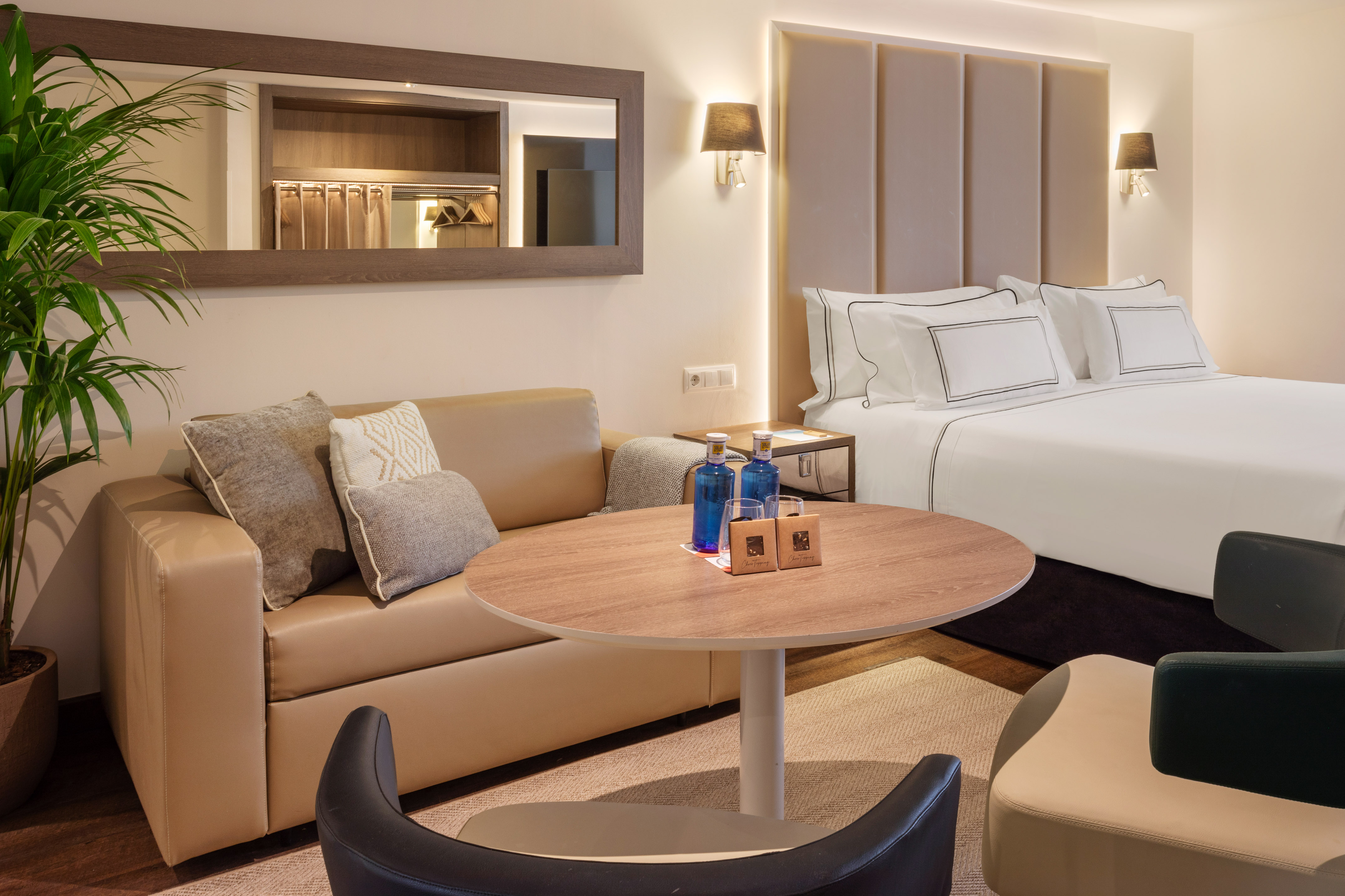 Modern hotel suite with large bed, beige sofa, round table, and bottled water.
