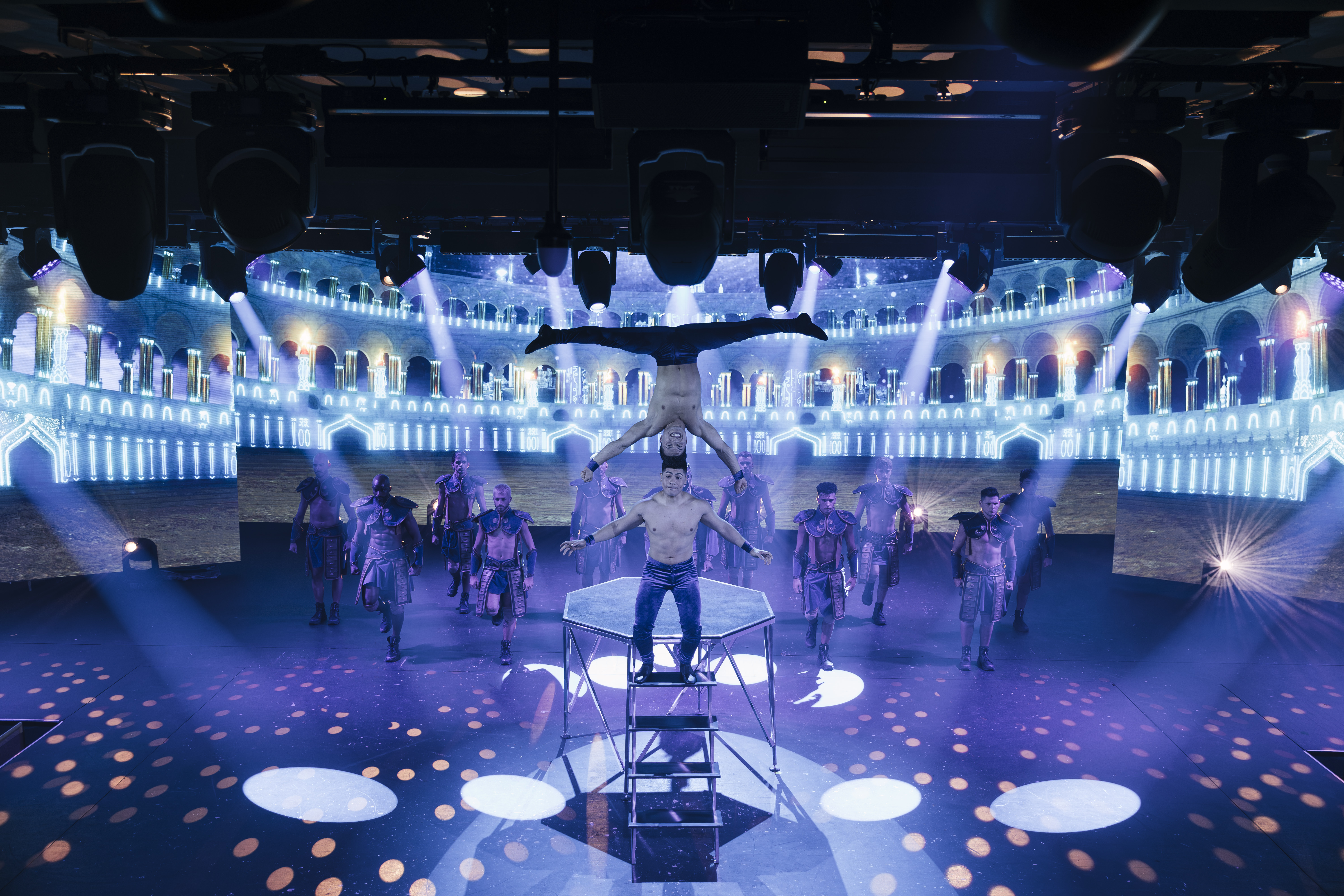 Acrobats balance in handstand above gladiator performers on a glowing stage.