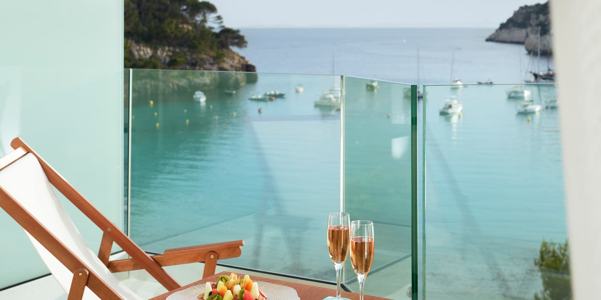 a table with a plate of fruit and wine glasses on a balcony overlooking a body of water