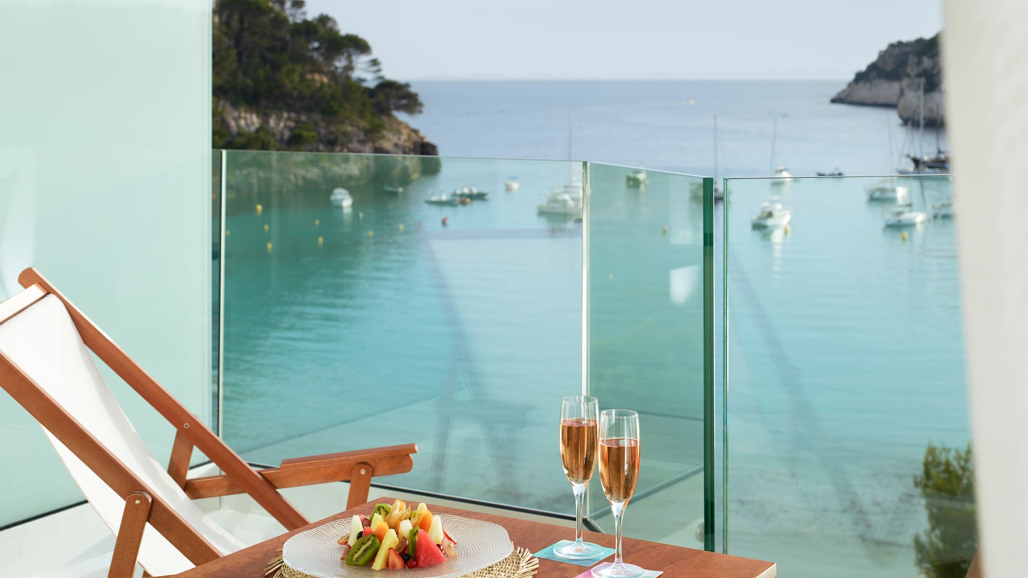 a table with a plate of fruit and wine glasses on a balcony overlooking a body of water