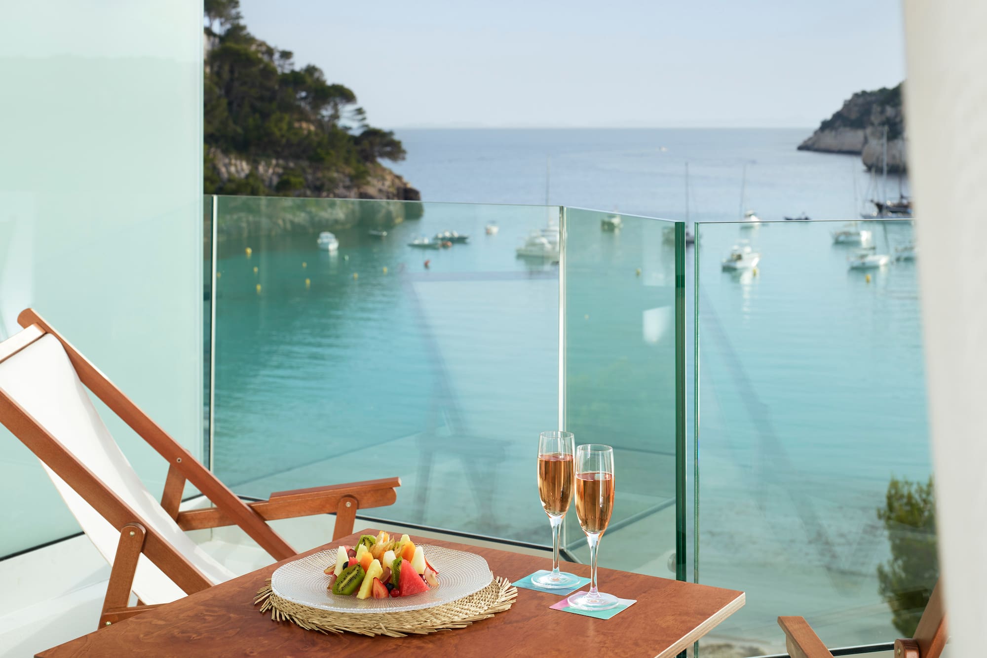 a table with a plate of fruit and wine glasses on a balcony overlooking a body of water