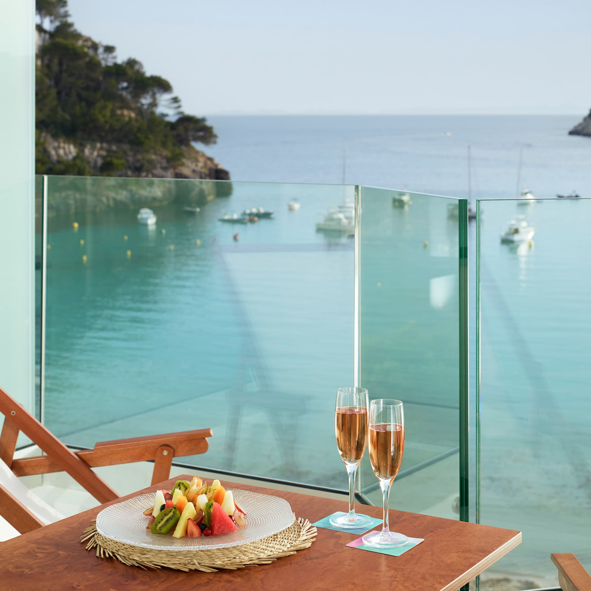 a table with a plate of fruit and wine glasses on a balcony overlooking a body of water