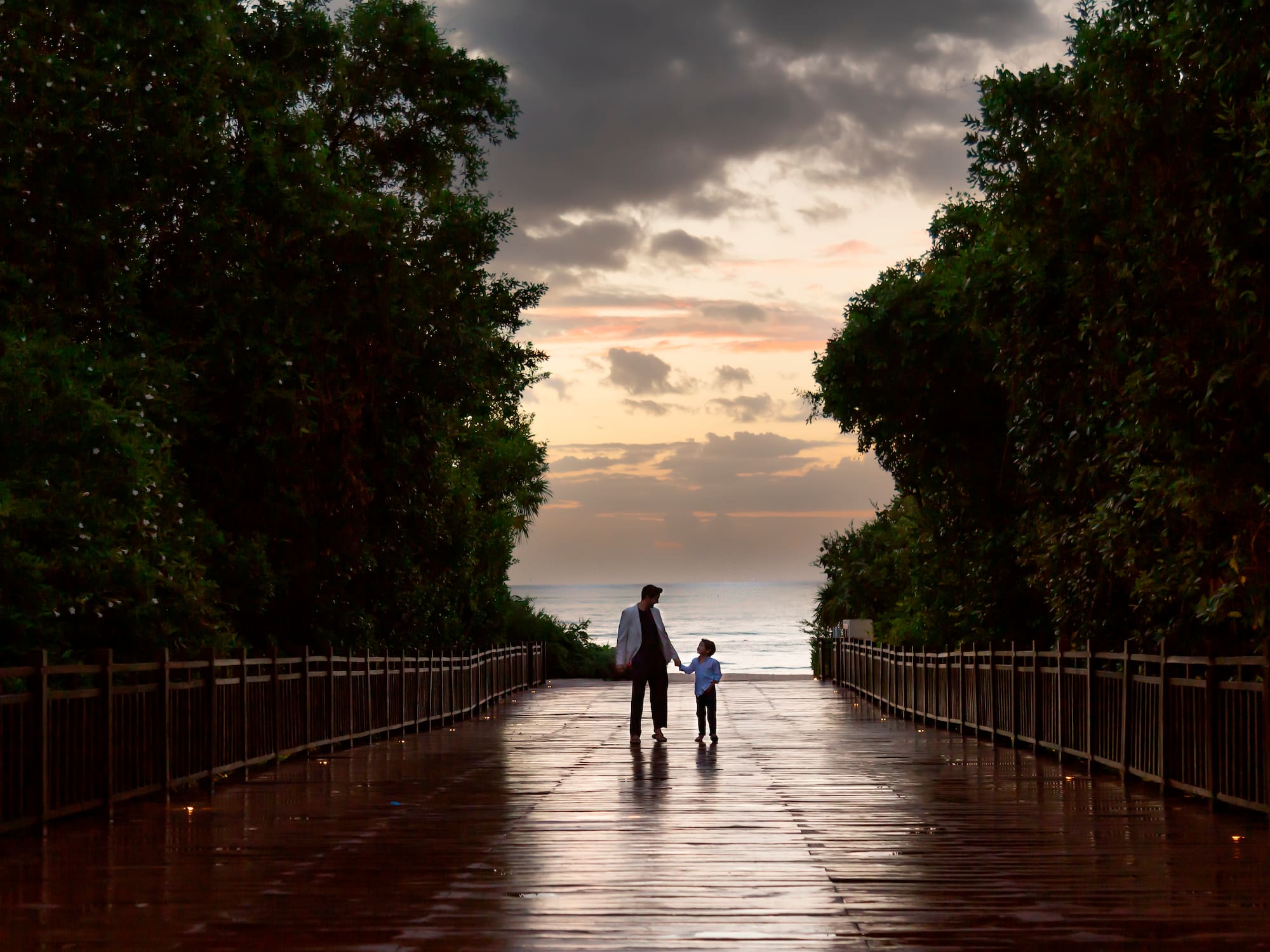 a man and child walking on a wet walkway with trees and water
