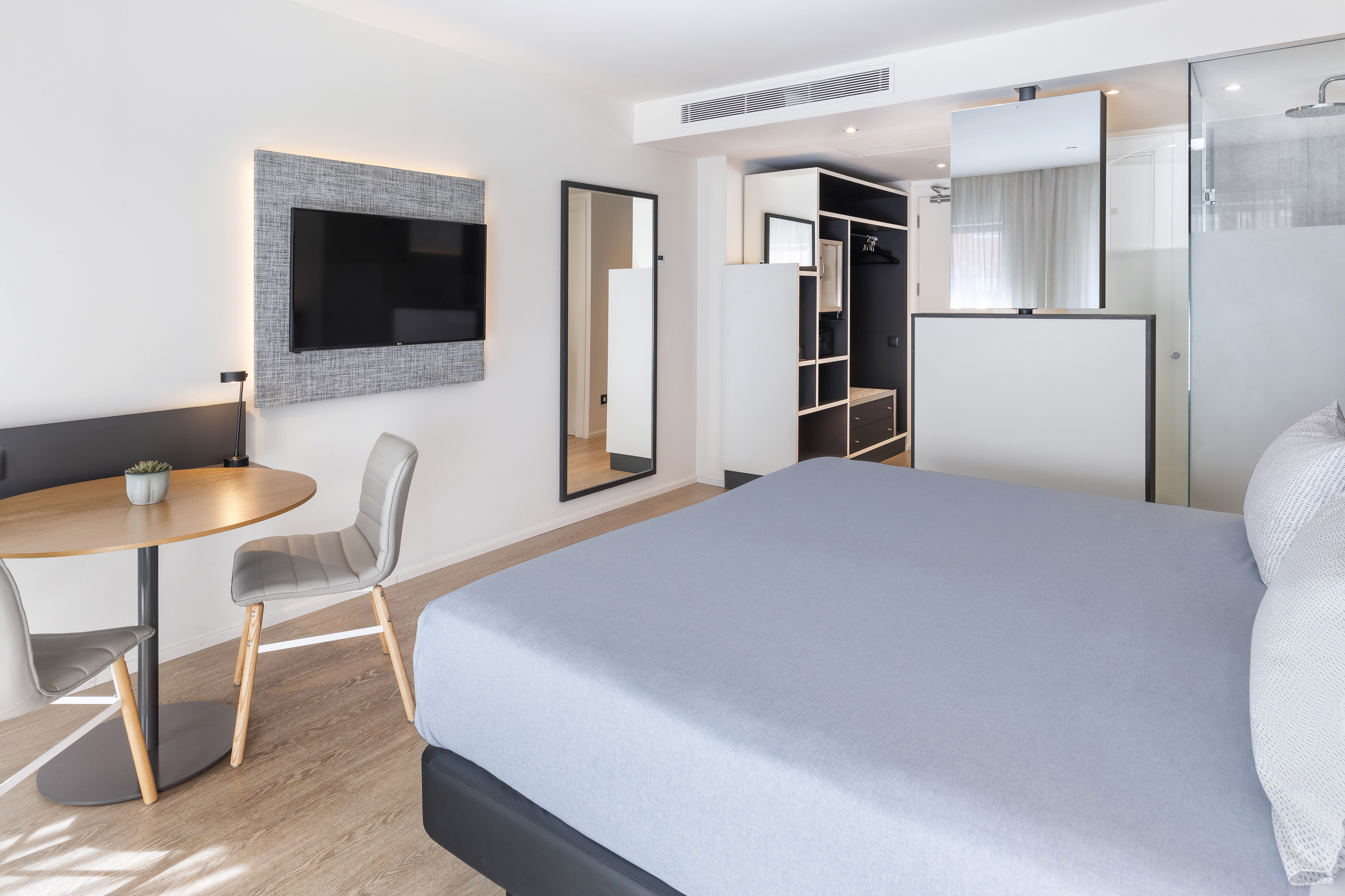 Stylish hotel bedroom with light blue bed, TV, wooden table, and glass shower.