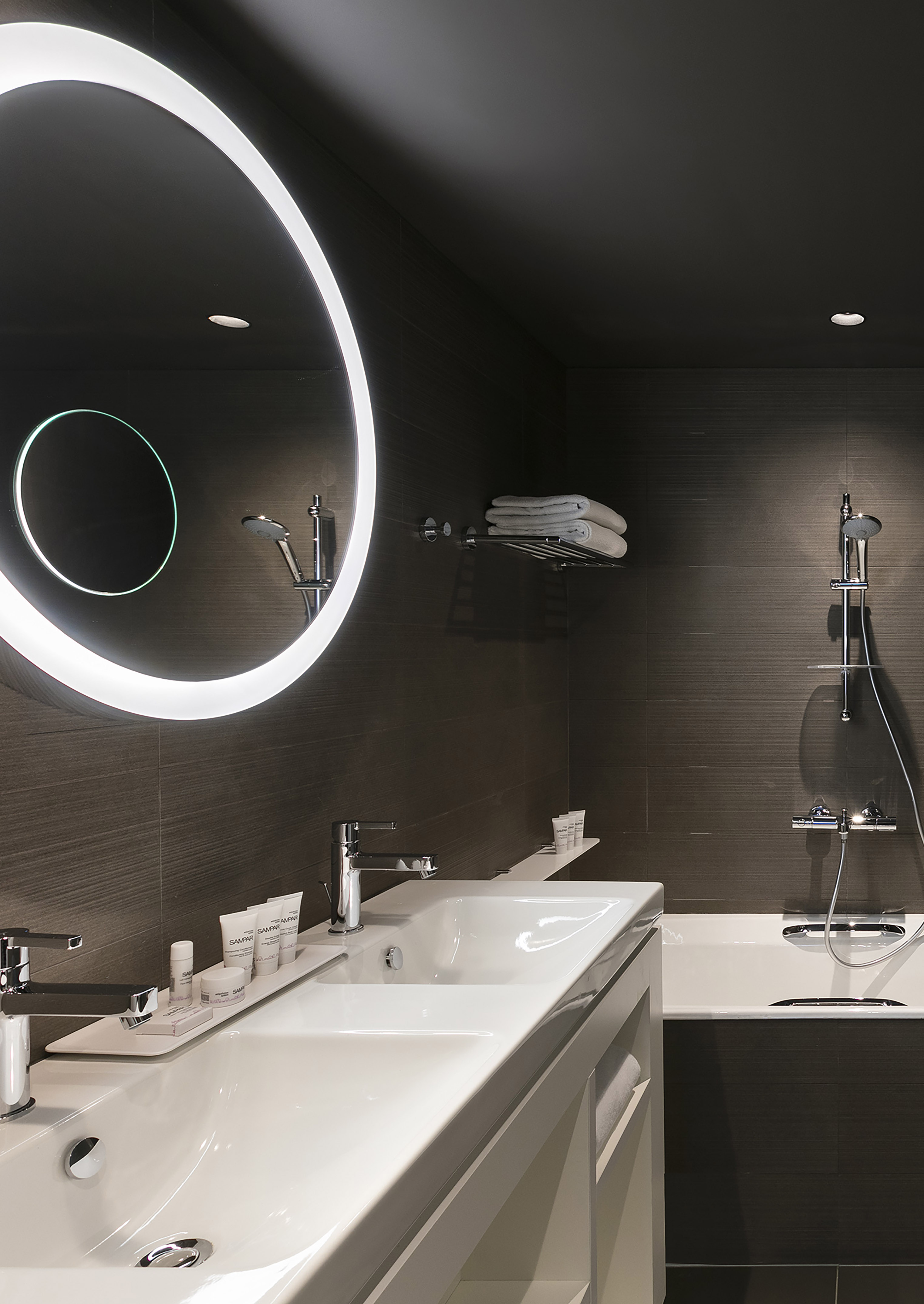 Modern hotel bathroom with double vanity, illuminated mirror, bathtub, and dark tiles.
