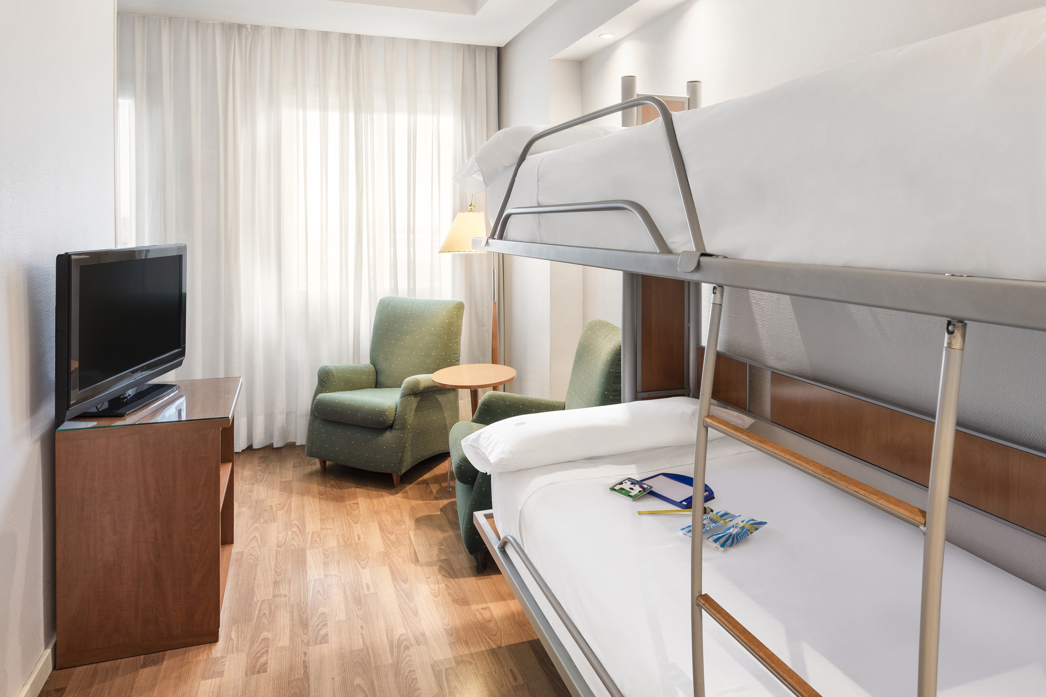 Hotel family room with metal bunk beds, television, and green armchairs.
