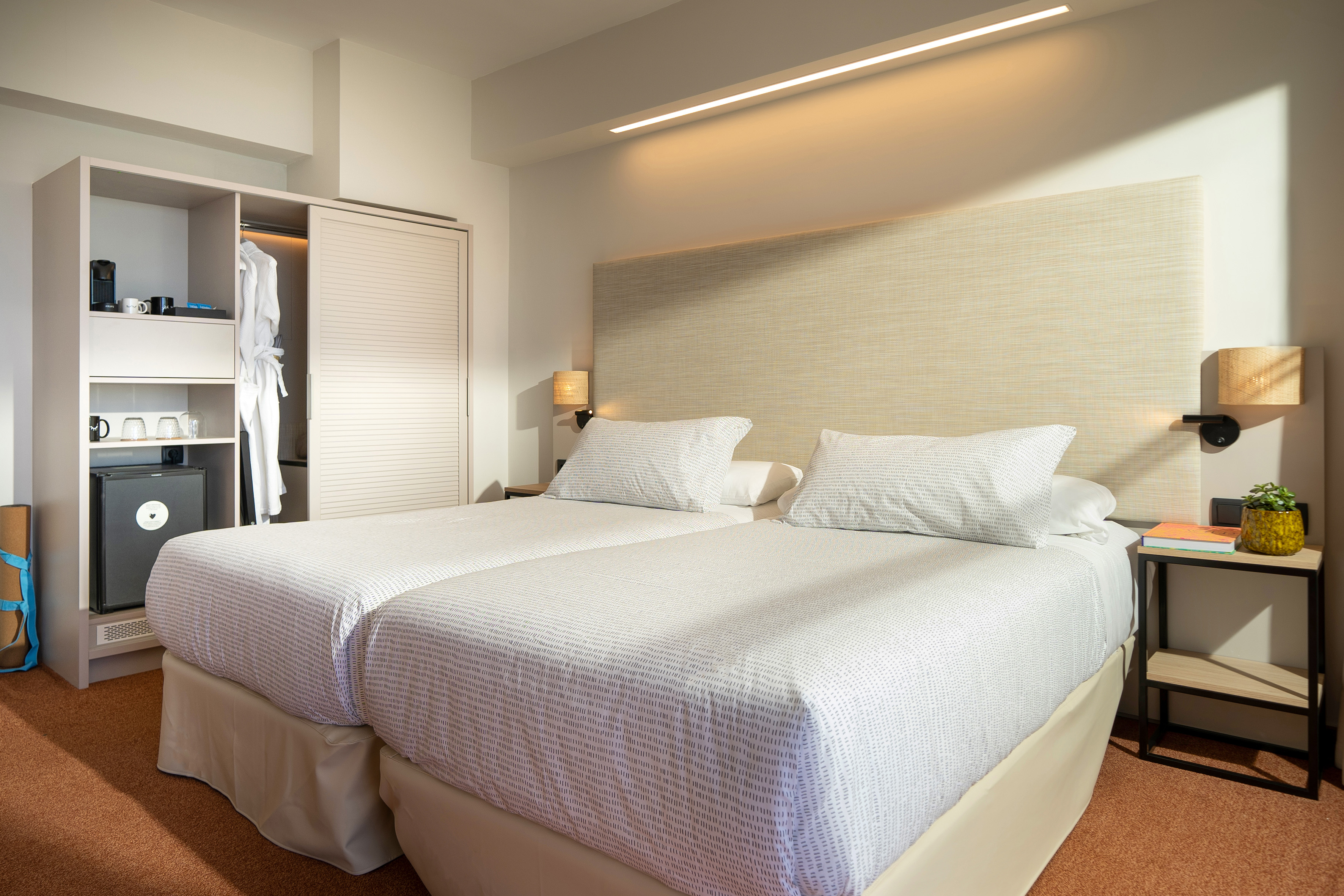 Modern hotel room with two twin beds, closet, mini-fridge, and stylish lighting.