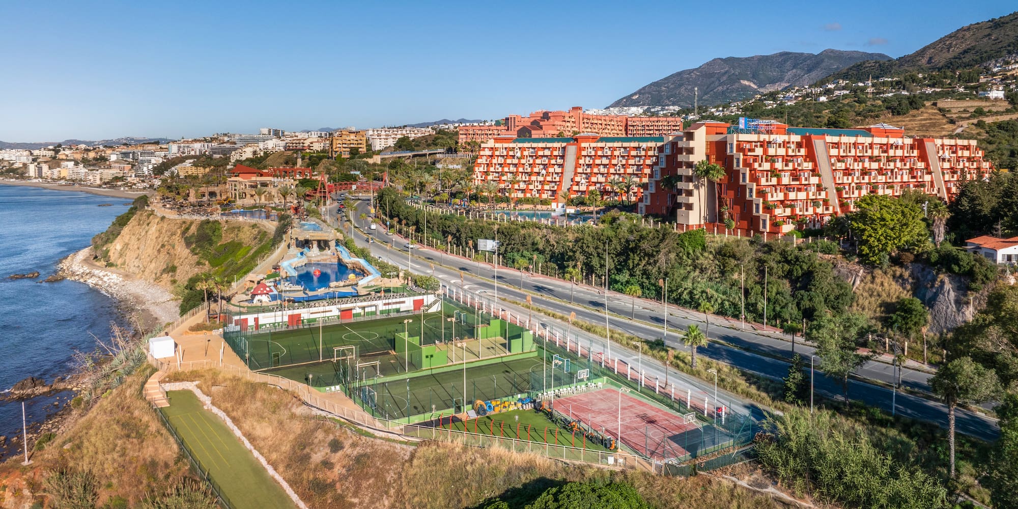 Coastal resort with orange hotels, sports courts, swimming pools, ocean, and mountains.