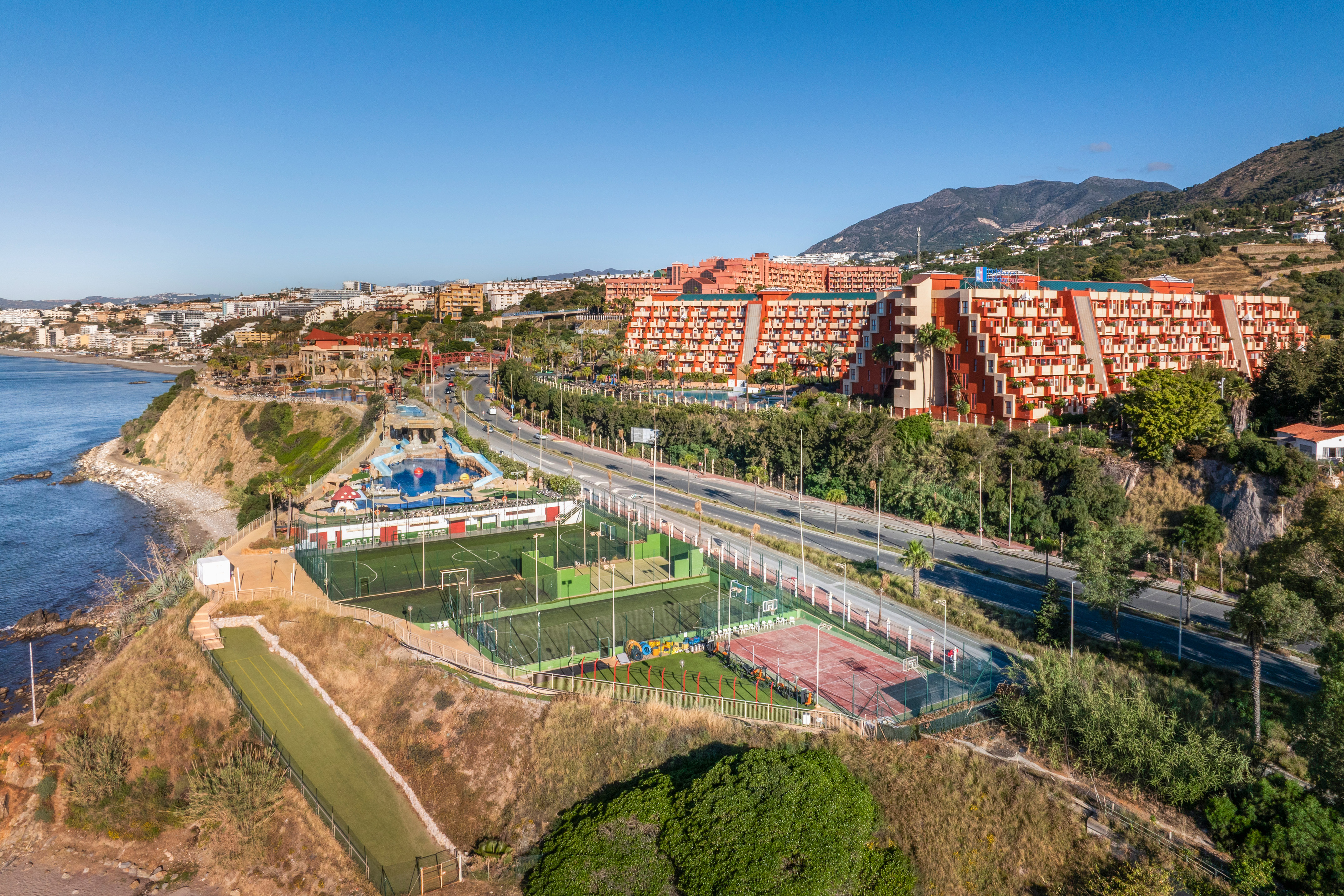 Coastal resort with orange hotels, sports courts, swimming pools, ocean, and mountains.