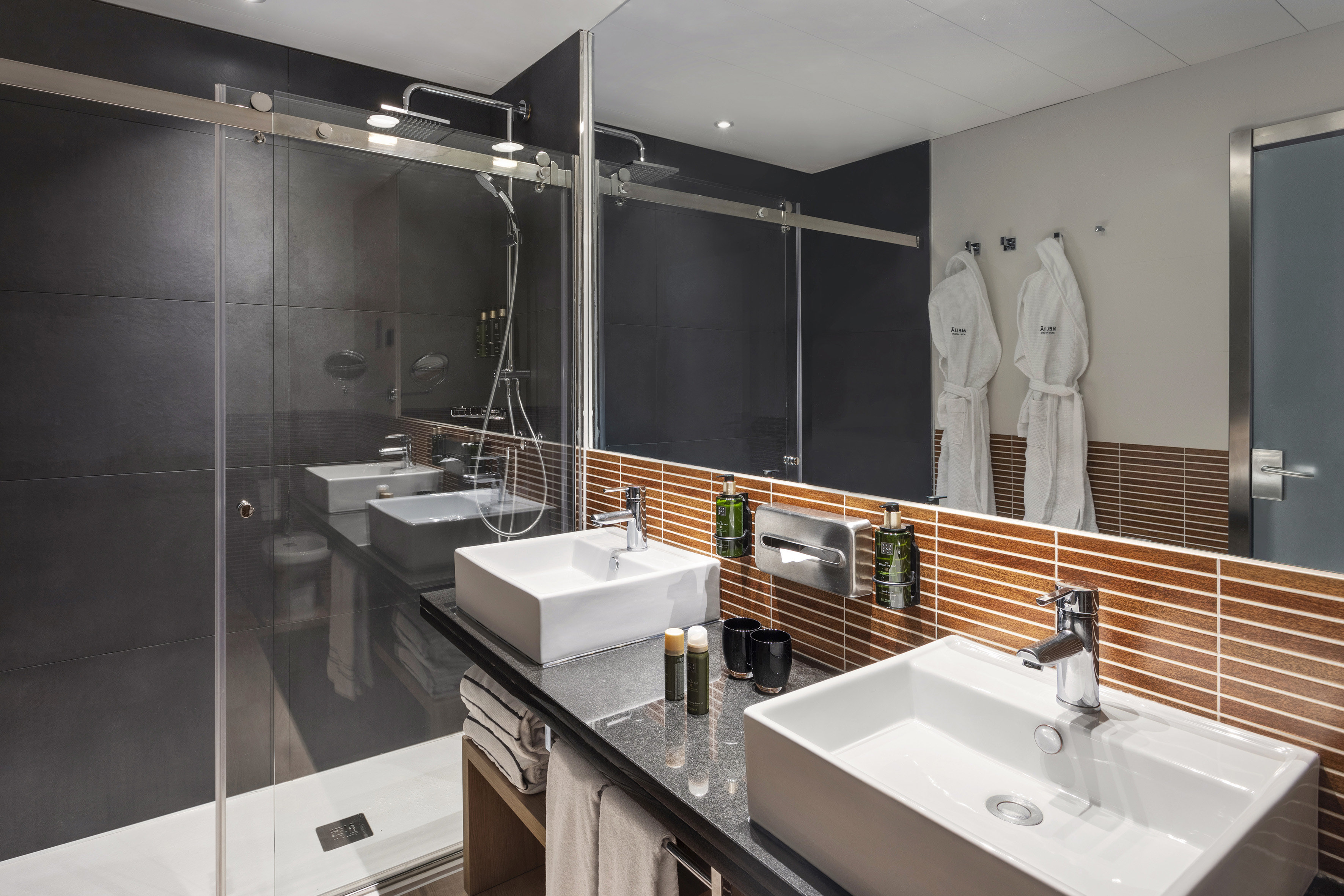 Modern hotel bathroom with double vanity, glass walk-in shower, and white robes.