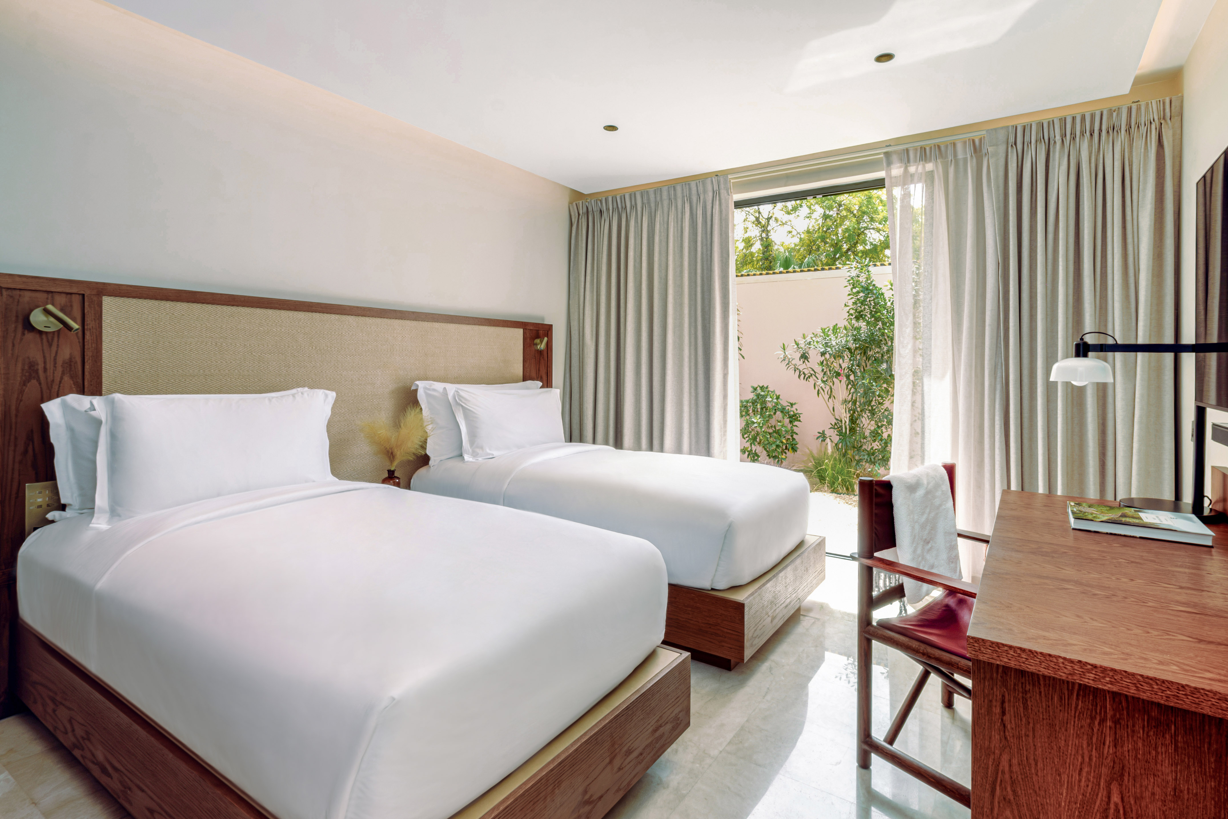 Bright hotel room with two single beds, wooden desk, and garden access.