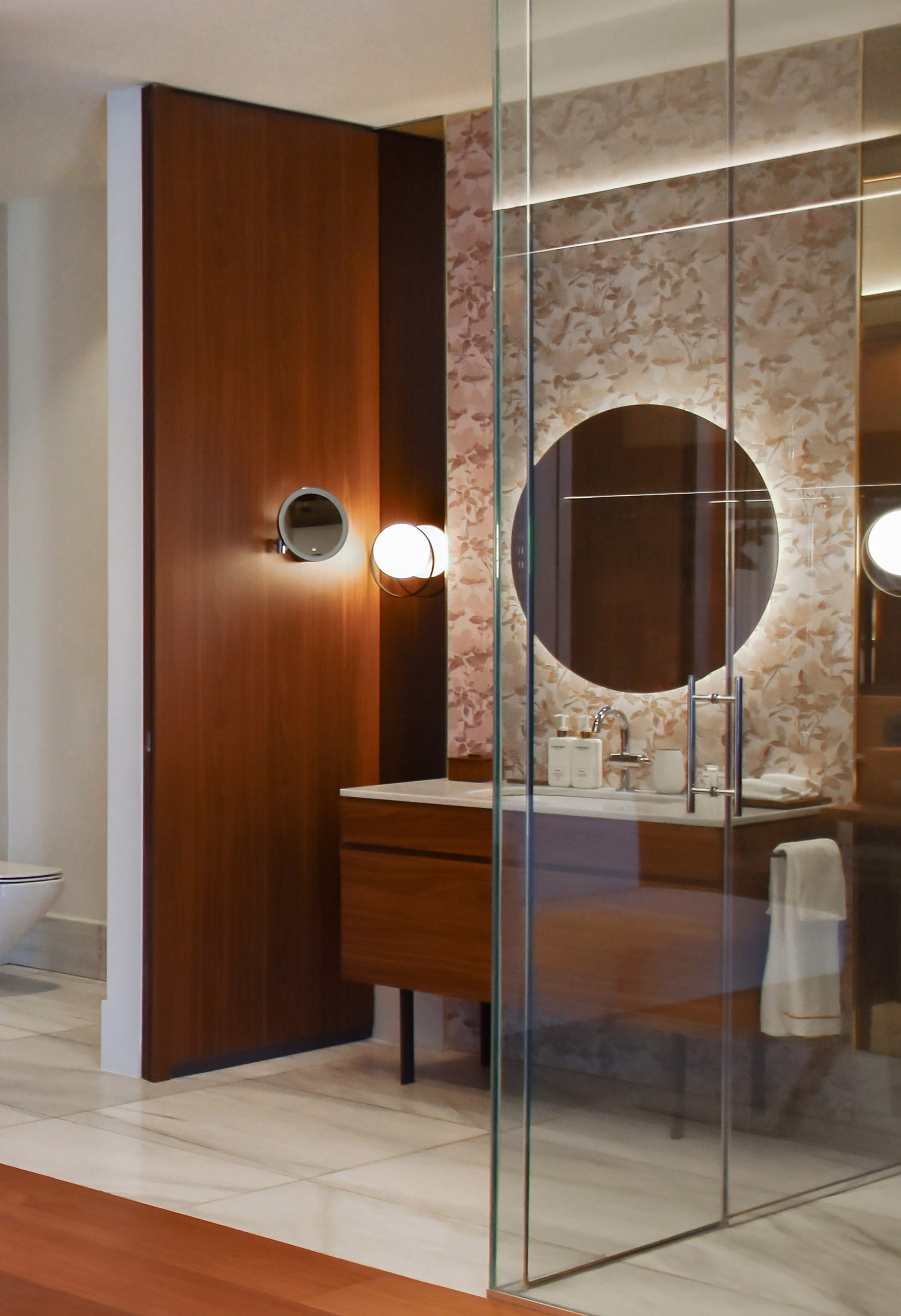 Sleek modern bathroom with wood vanity, round backlit mirror, and glass shower.