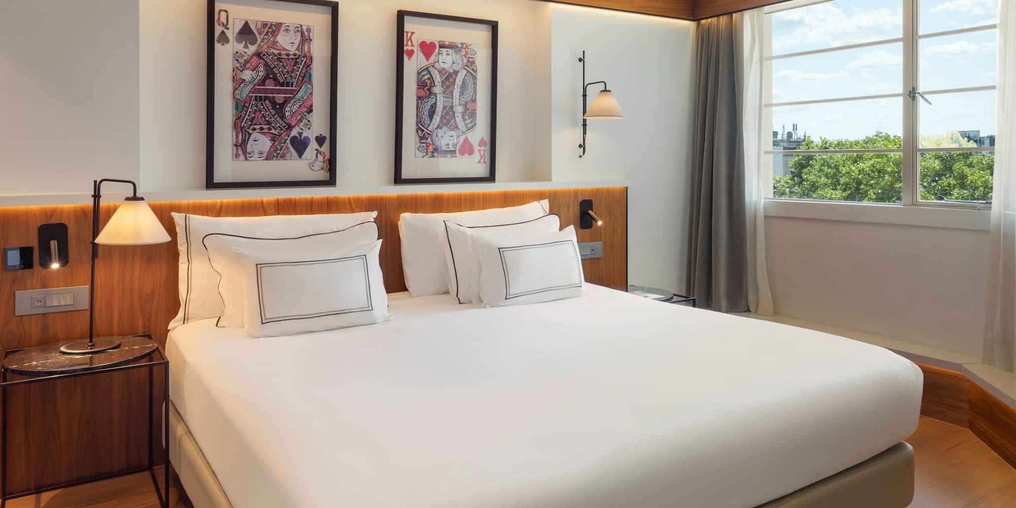 Modern hotel bedroom with white bed, playing card art, and bright window view.
