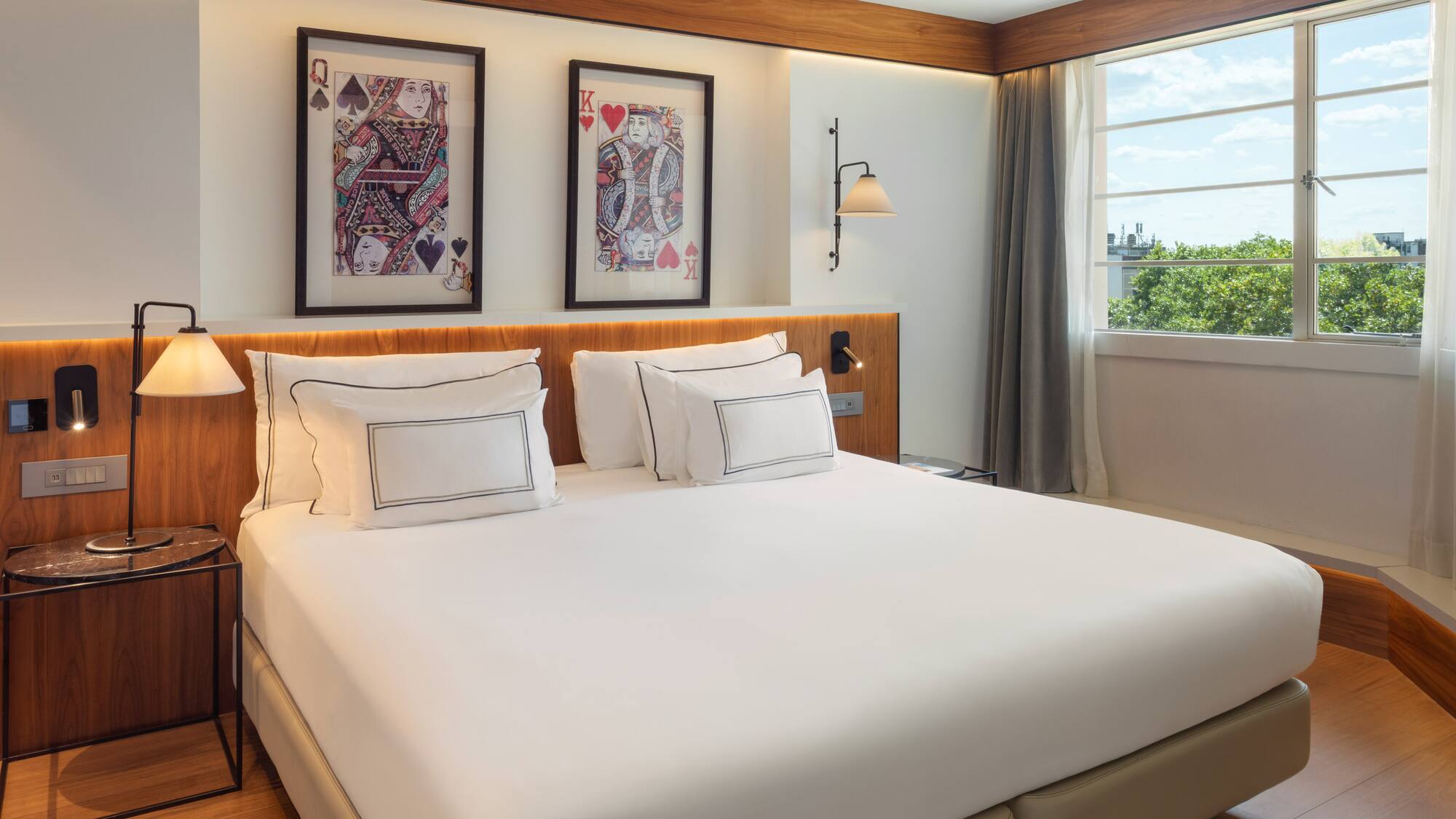 Contemporary hotel bedroom: white bed, card art, wooden details, and bright window view.