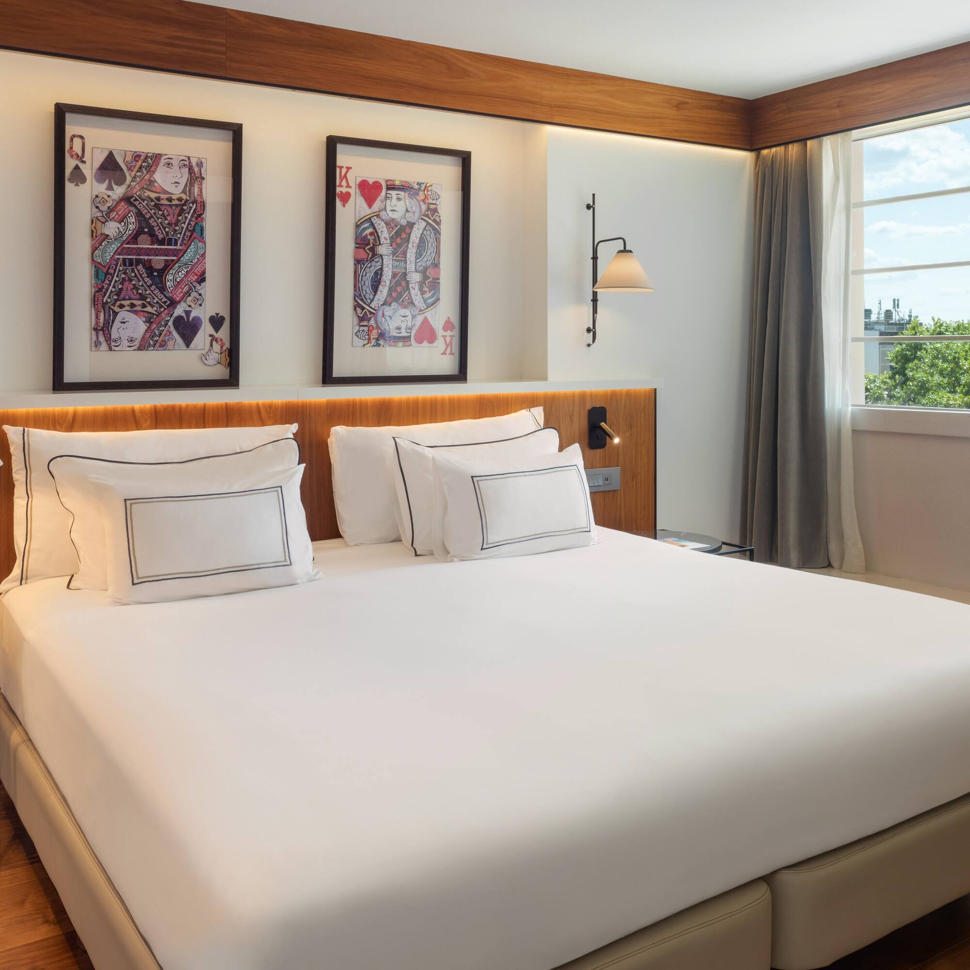 Contemporary hotel bedroom: white bed, card art, wooden details, and bright window view.