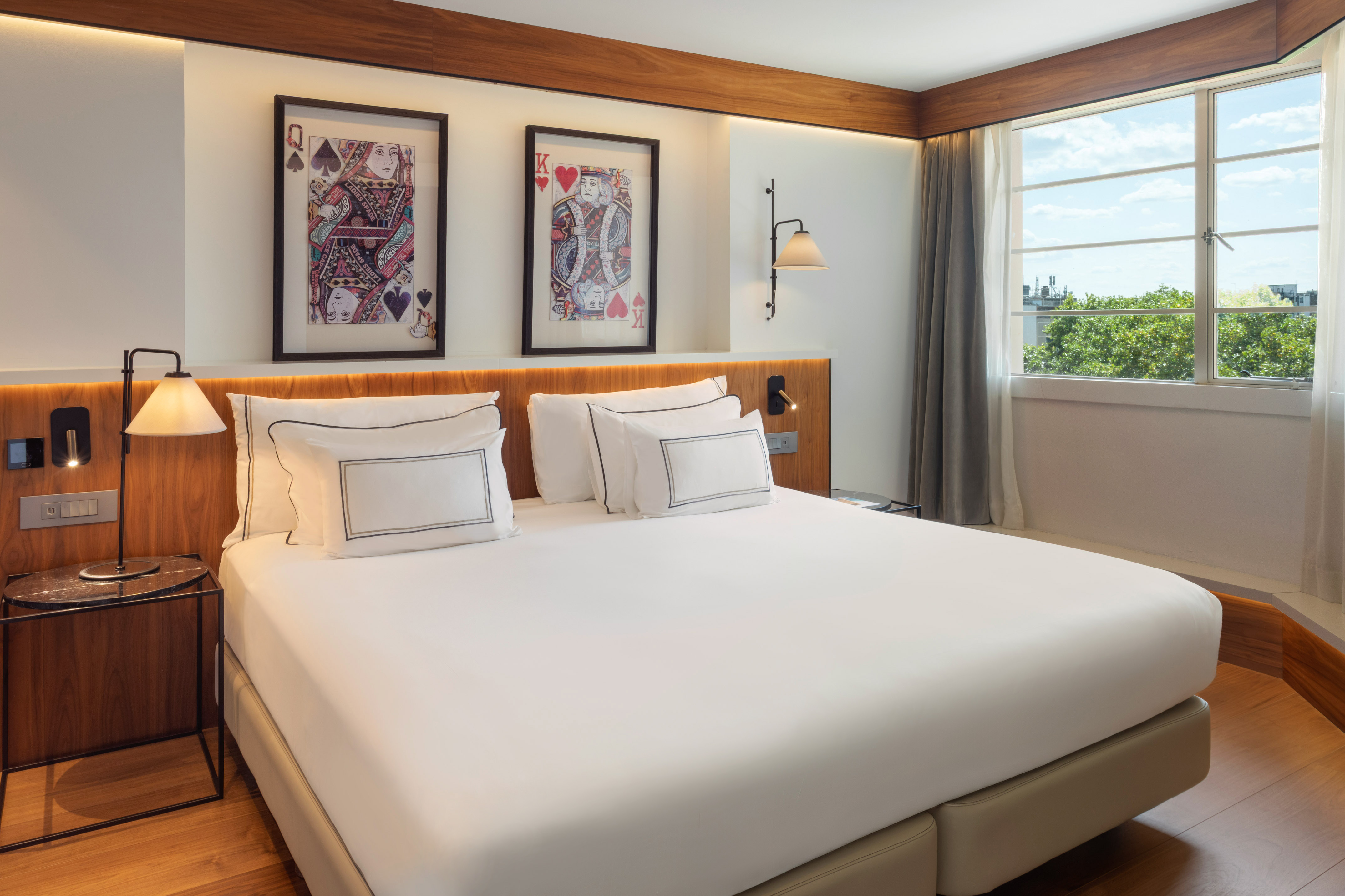 Contemporary hotel bedroom: white bed, card art, wooden details, and bright window view.
