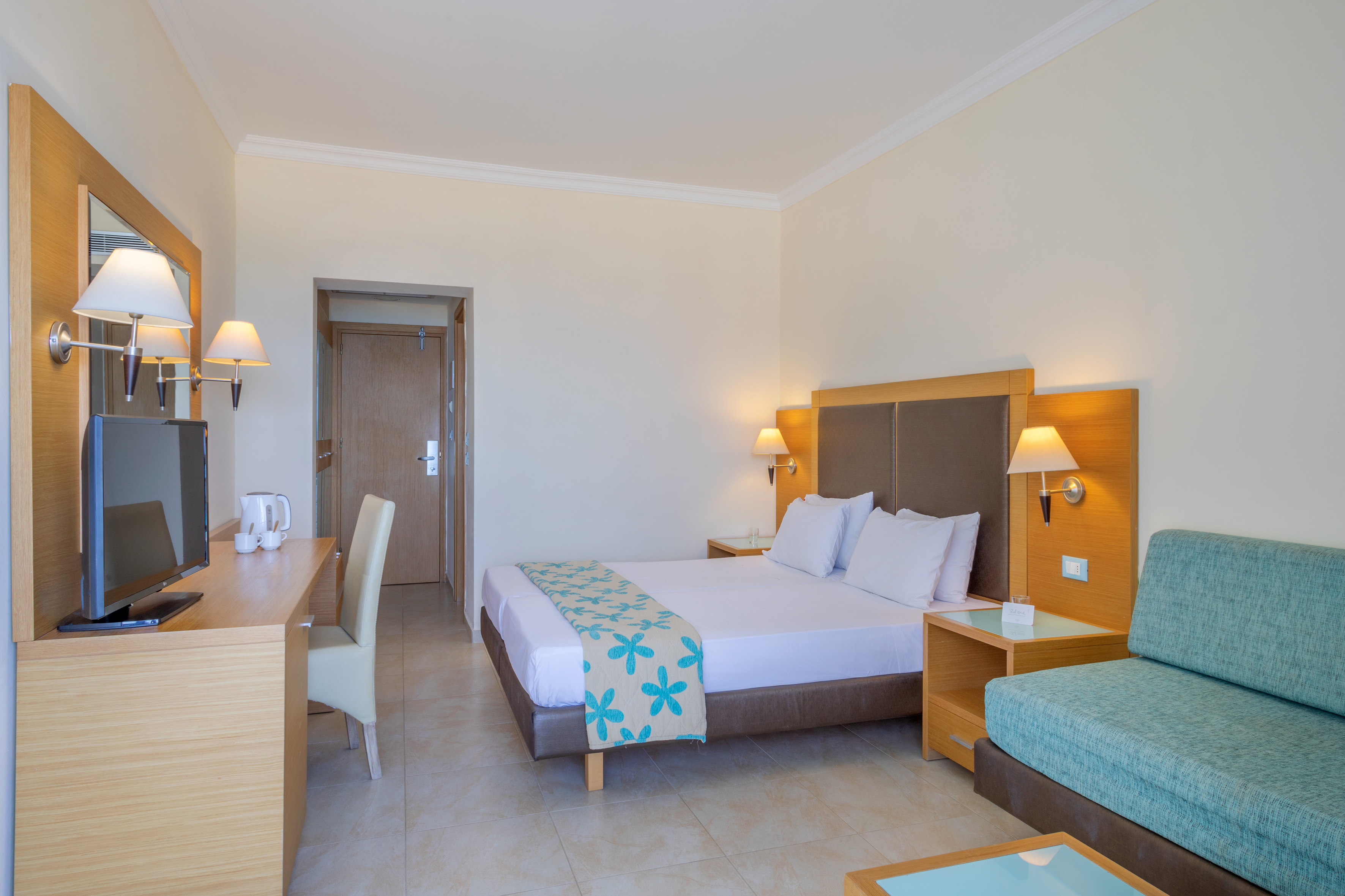 Bright hotel room with bed, teal sofa, wooden desk, and TV.