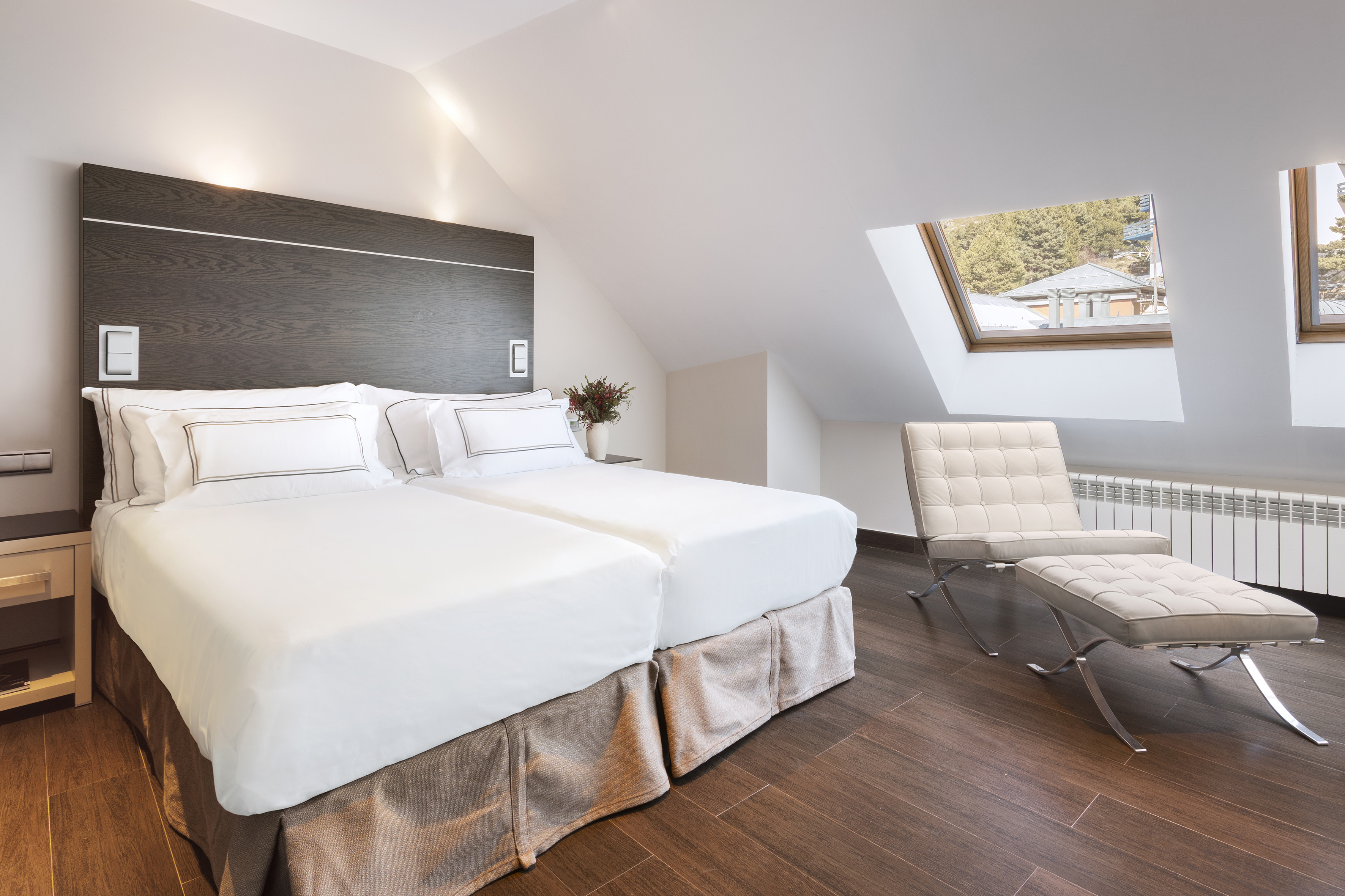 Bright loft hotel bedroom with two beds, skylight windows, and modern armchair.