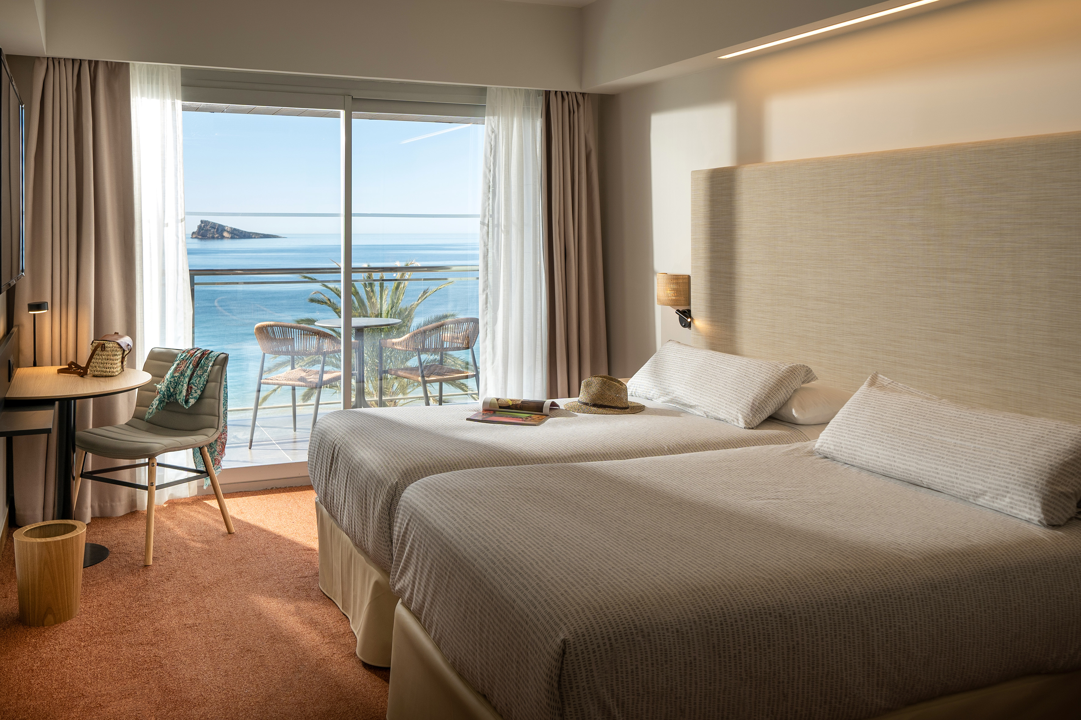 Bright modern hotel room with twin beds, balcony sea view, and distant island.