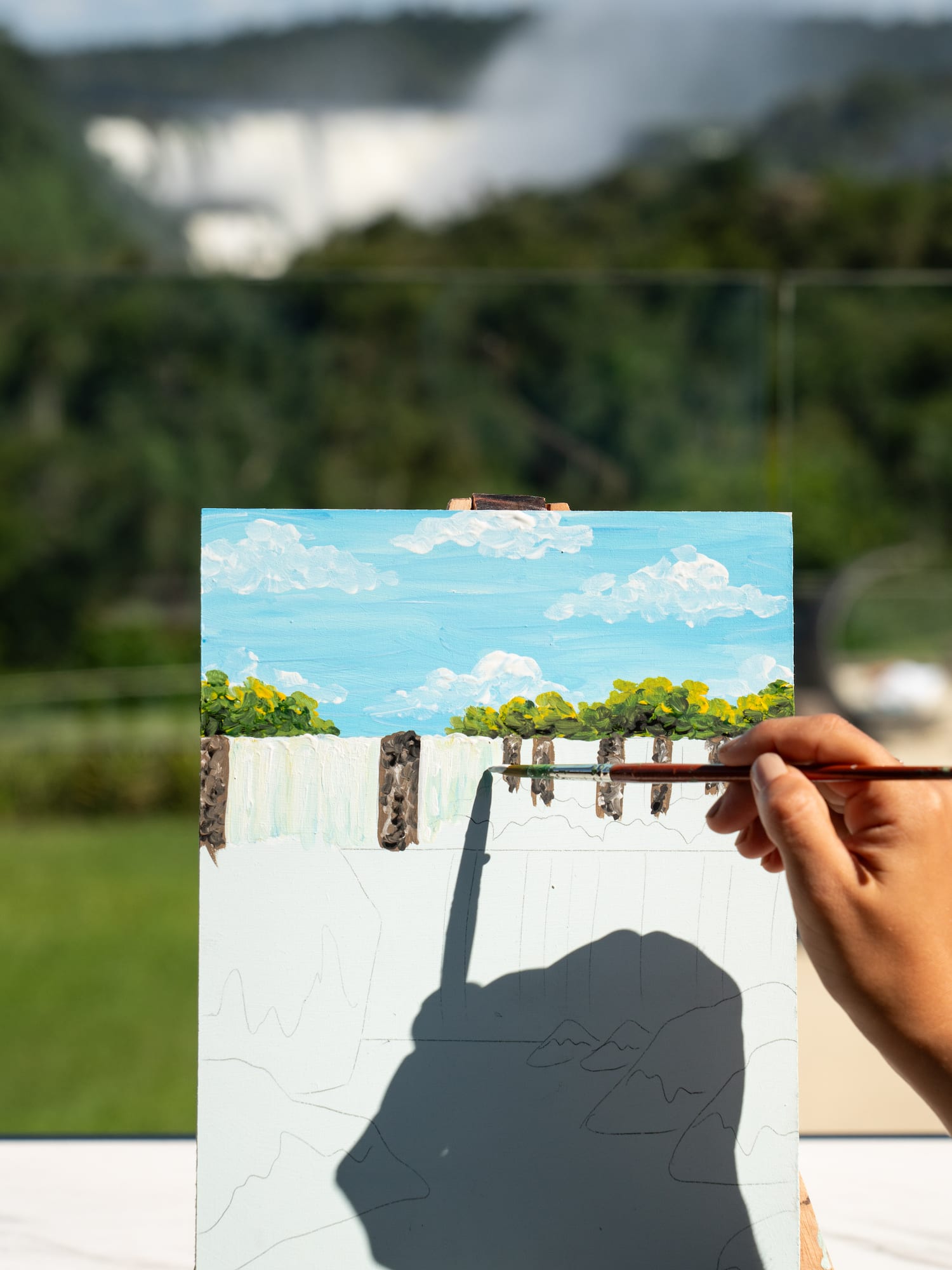 Artist's hand paints waterfall on easel, blurred real falls in the background.