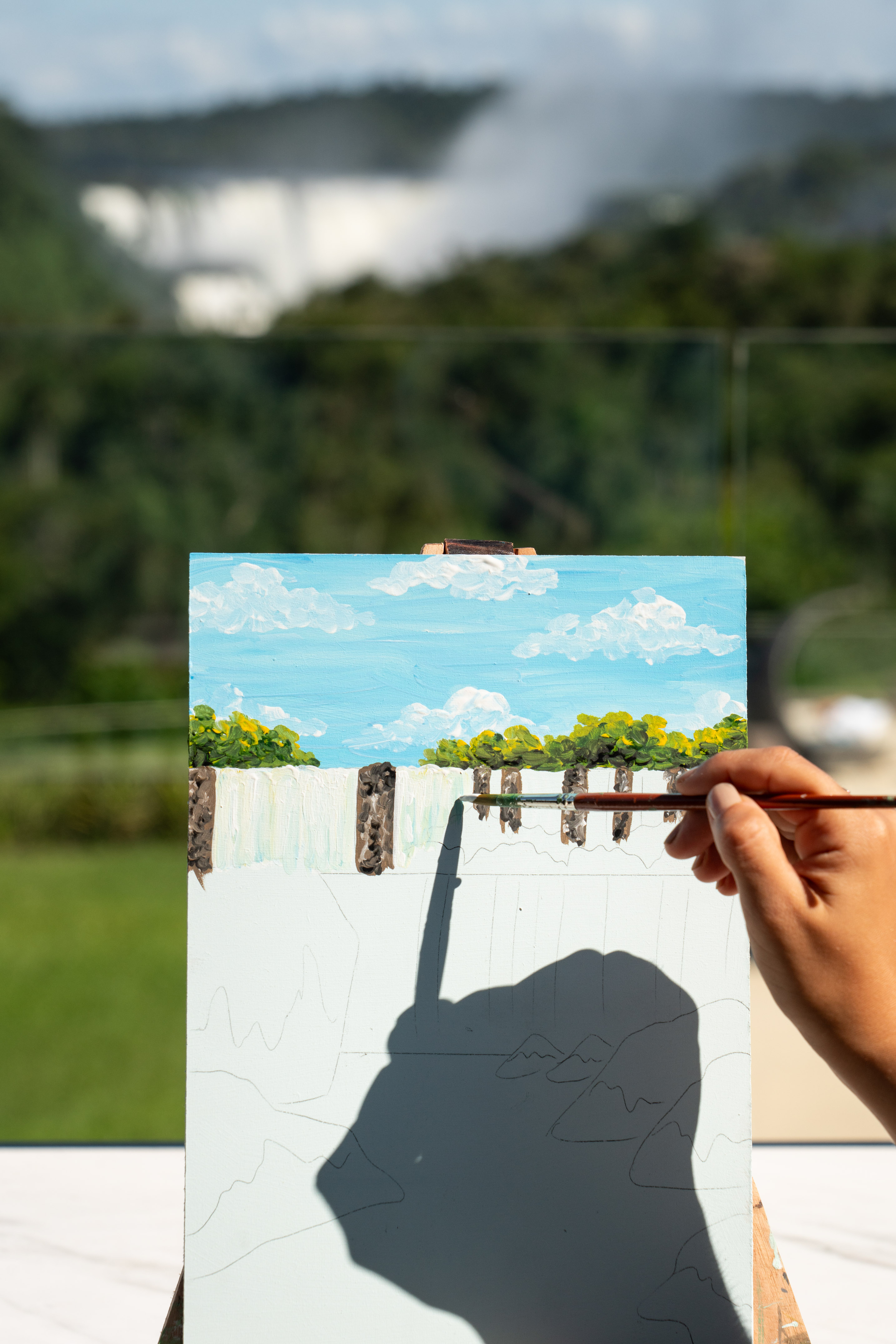 Artist's hand paints waterfall on easel, blurred real falls in the background.