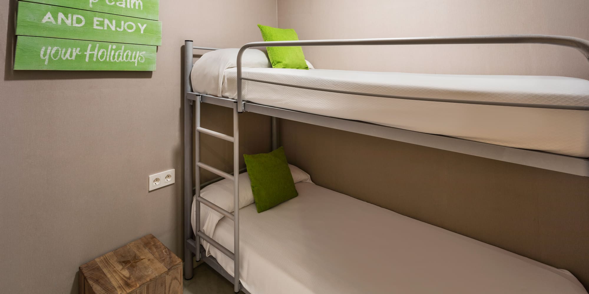 Hostel room with metal bunk bed, green pillows, and "enjoy your holidays" sign.