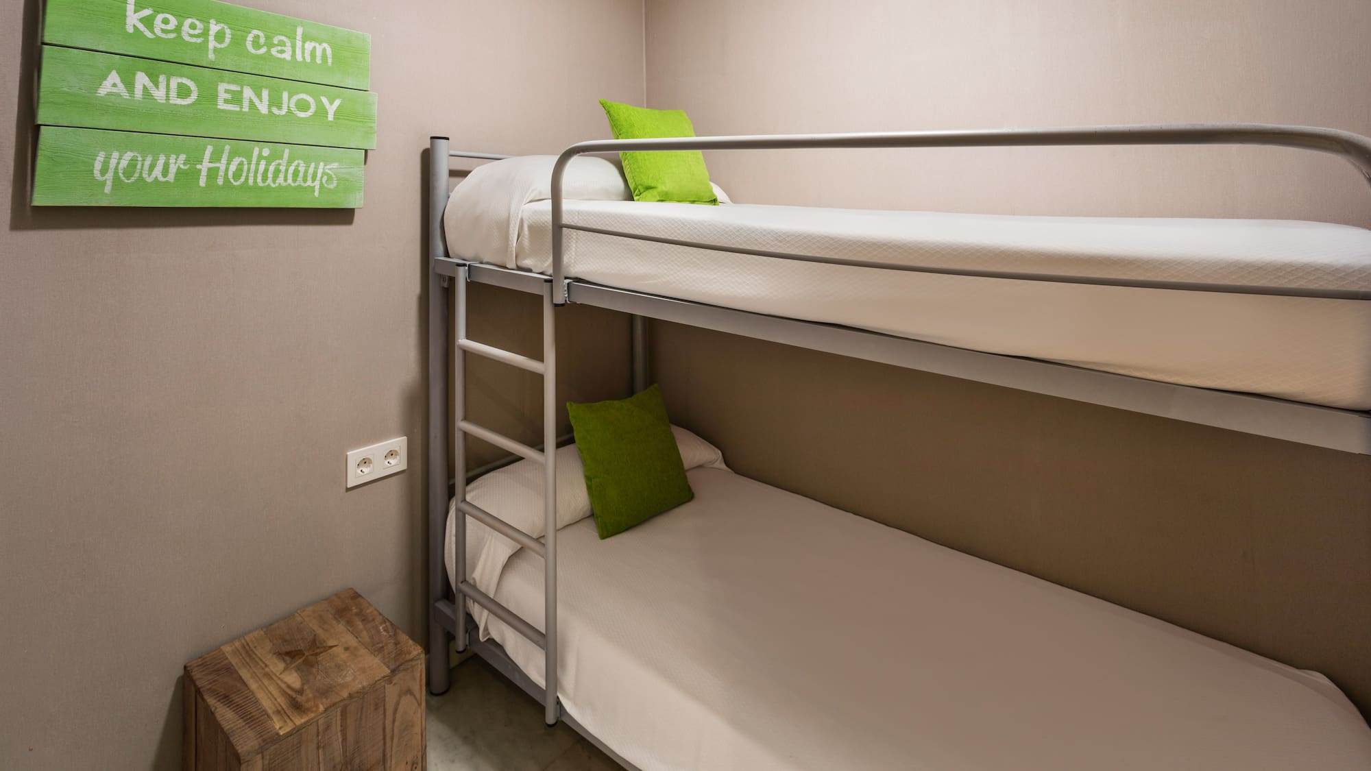 Hostel room with metal bunk bed, green pillows, and "enjoy your holidays" sign.