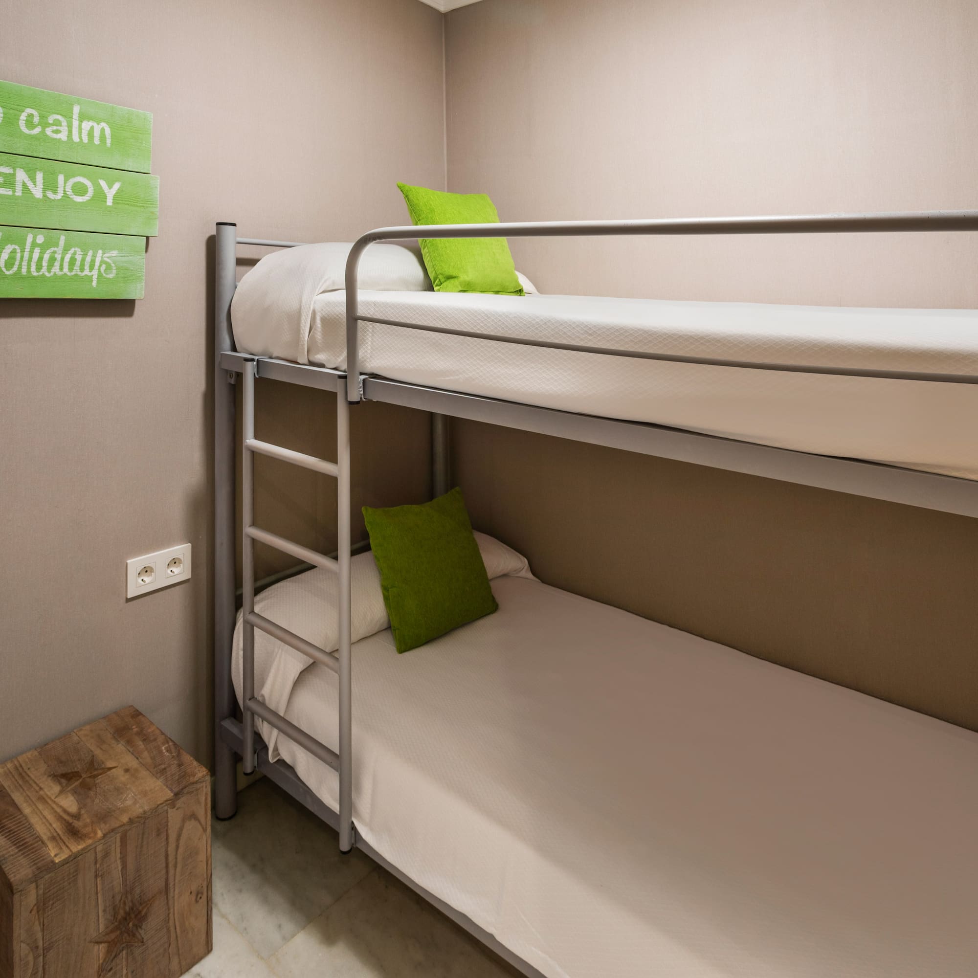 Hostel room with metal bunk bed, green pillows, and "enjoy your holidays" sign.
