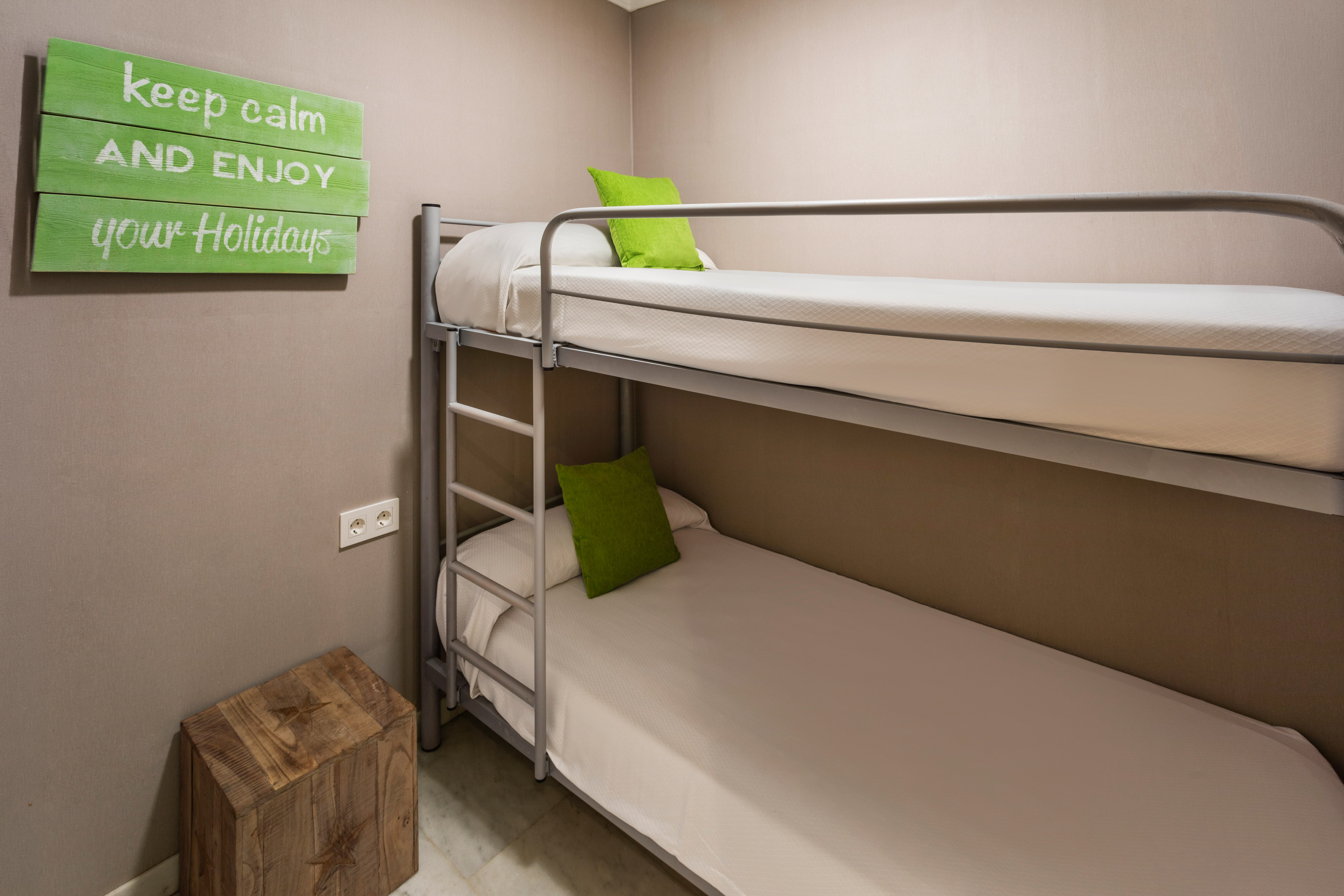 Hostel room with metal bunk bed, green pillows, and "enjoy your holidays" sign.