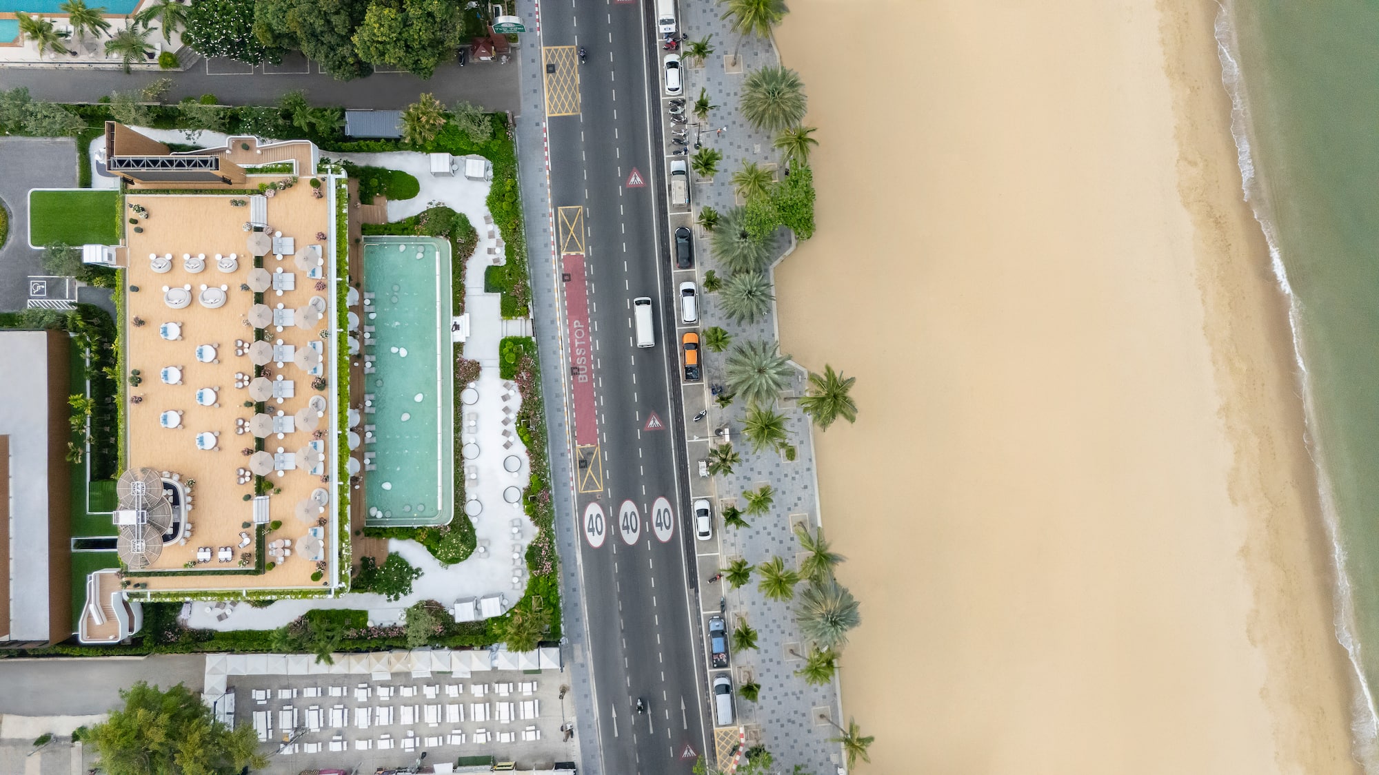 Aerial view: hotel resort, swimming pool, coastal road, palm trees, sandy beach, ocean.