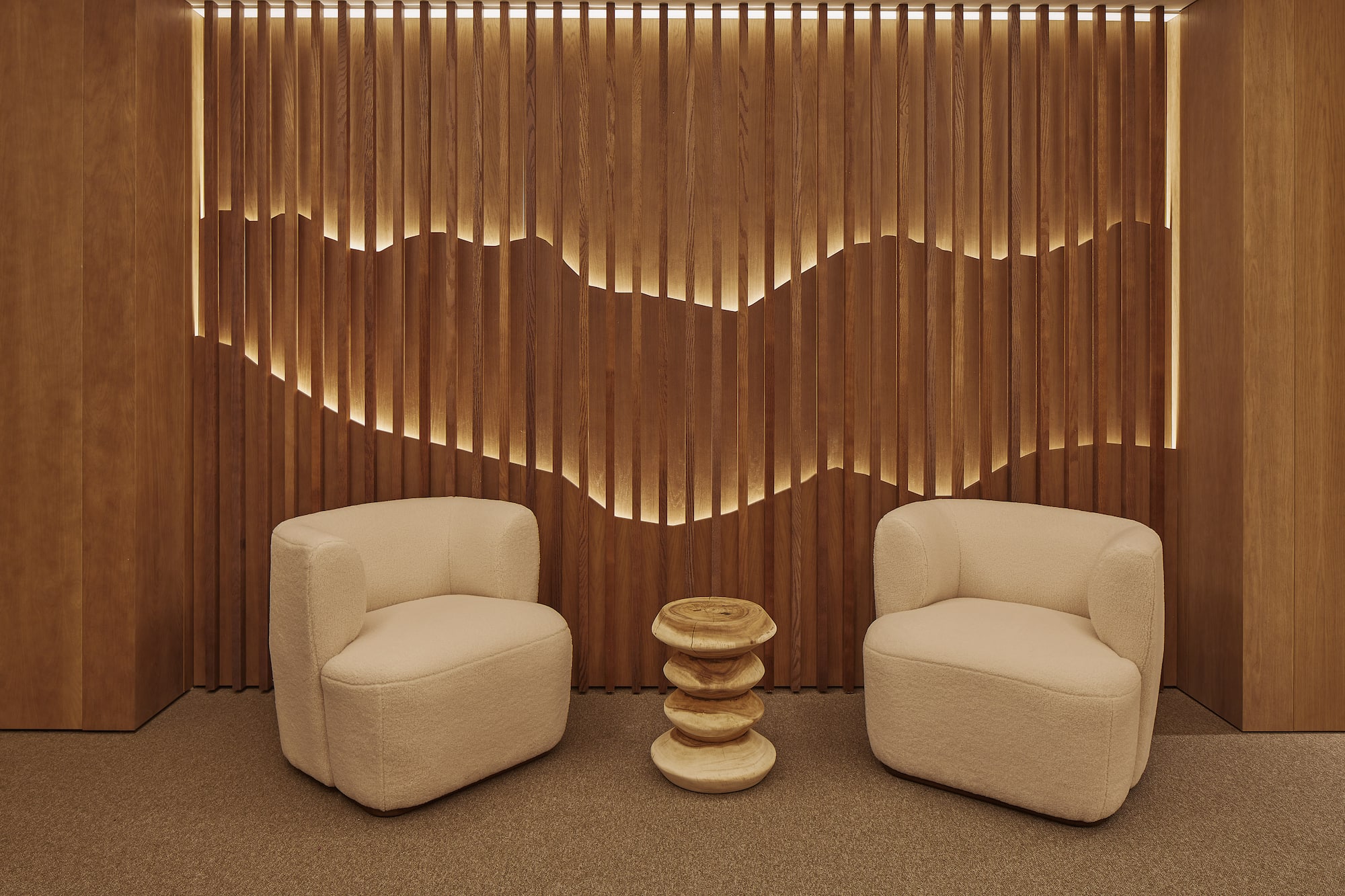 Cream lounge chairs, wooden stool, and backlit wavy slatted wood wall.