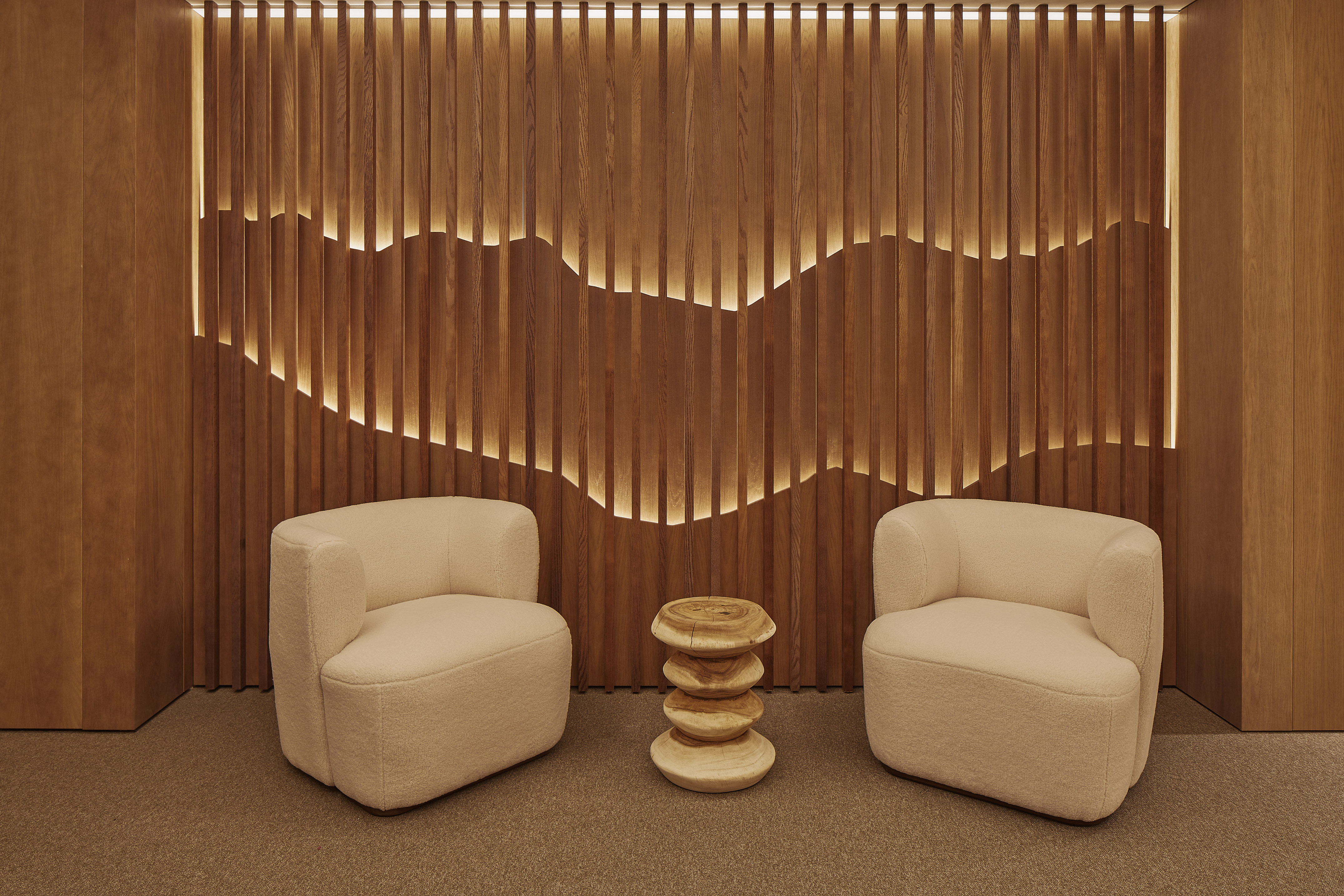 Cream lounge chairs, wooden stool, and backlit wavy slatted wood wall.