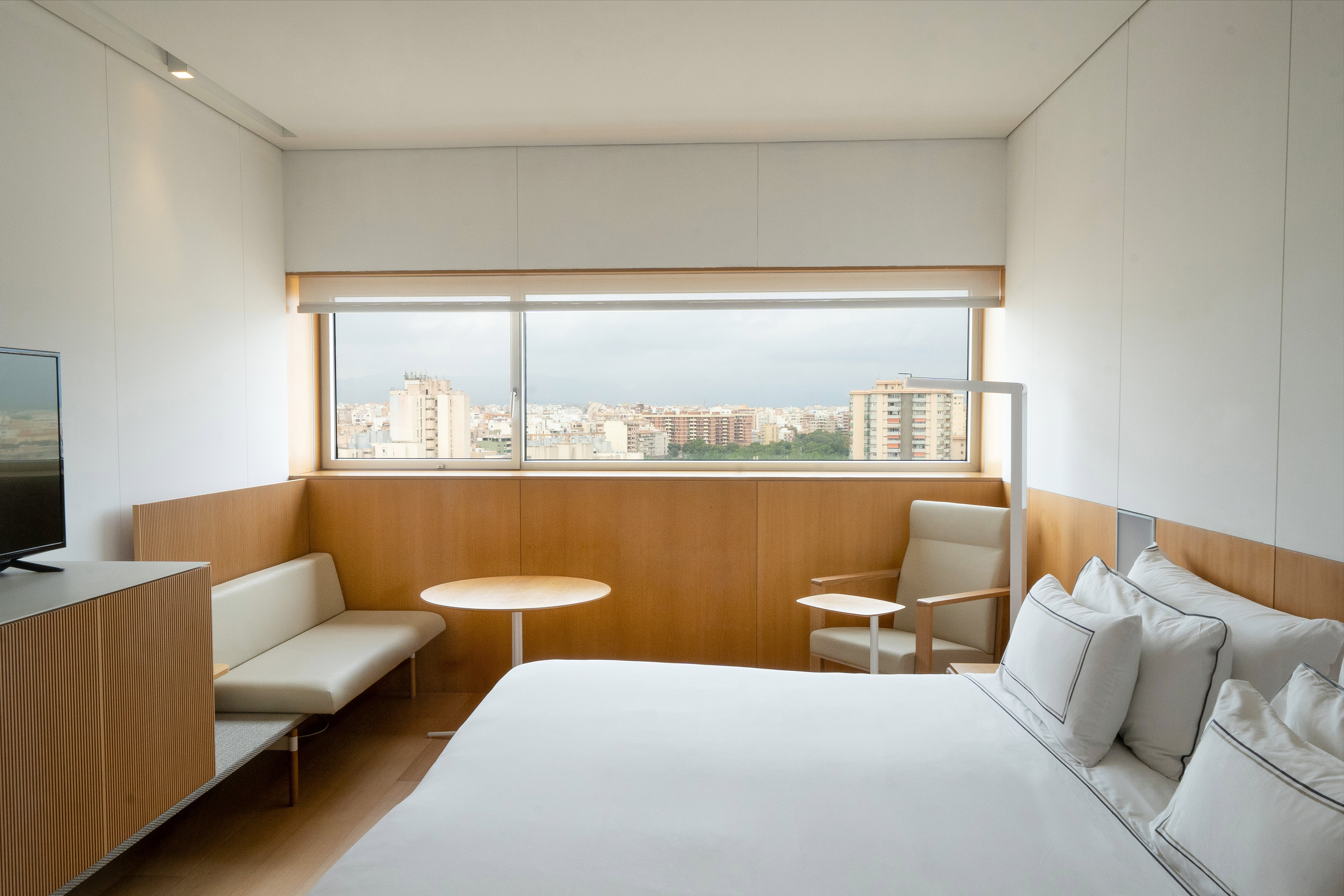 Modern hotel room with city view, white bed, wooden accents, and seating.