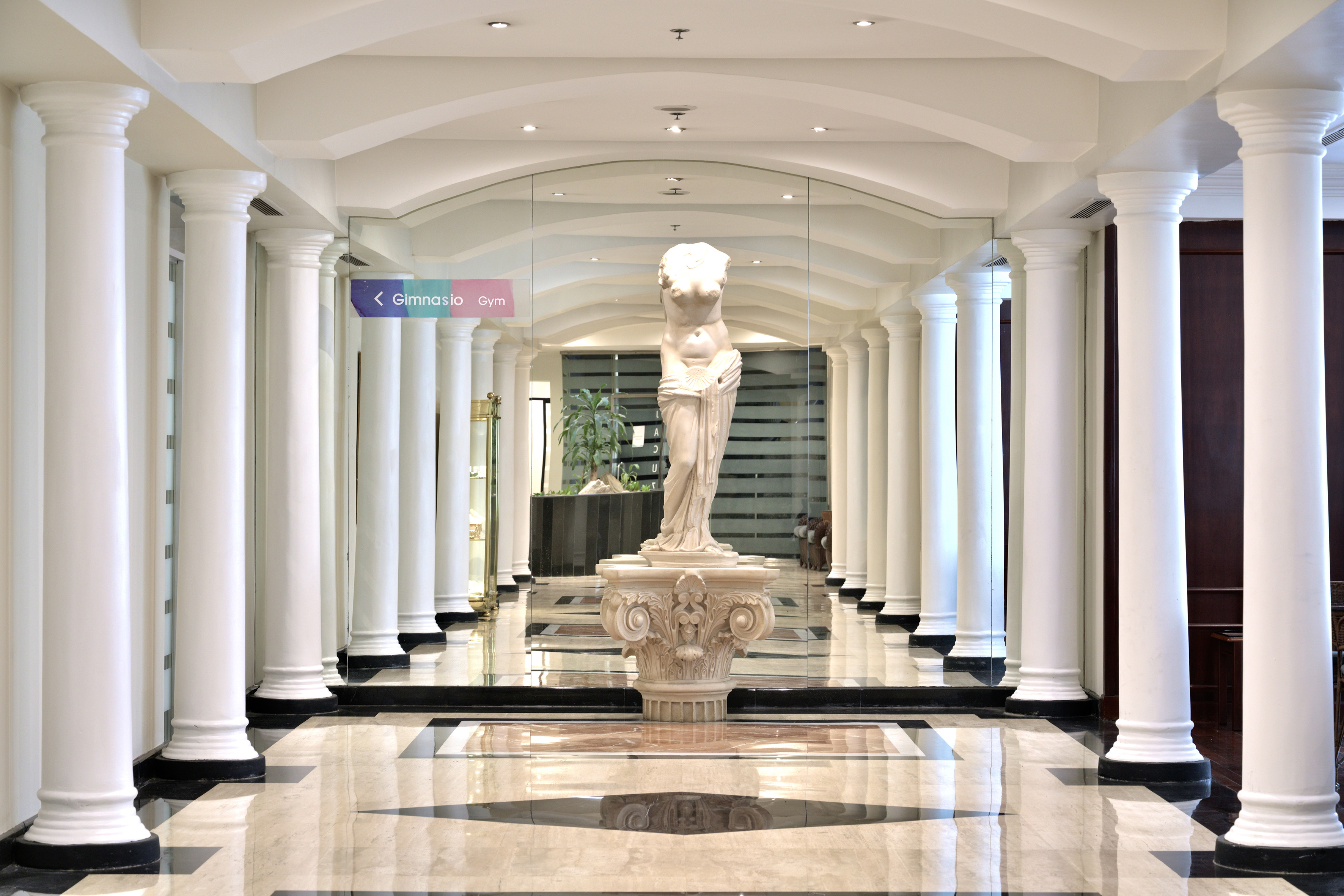 White columned hallway with marble statue, polished floor, and gym entrance sign.