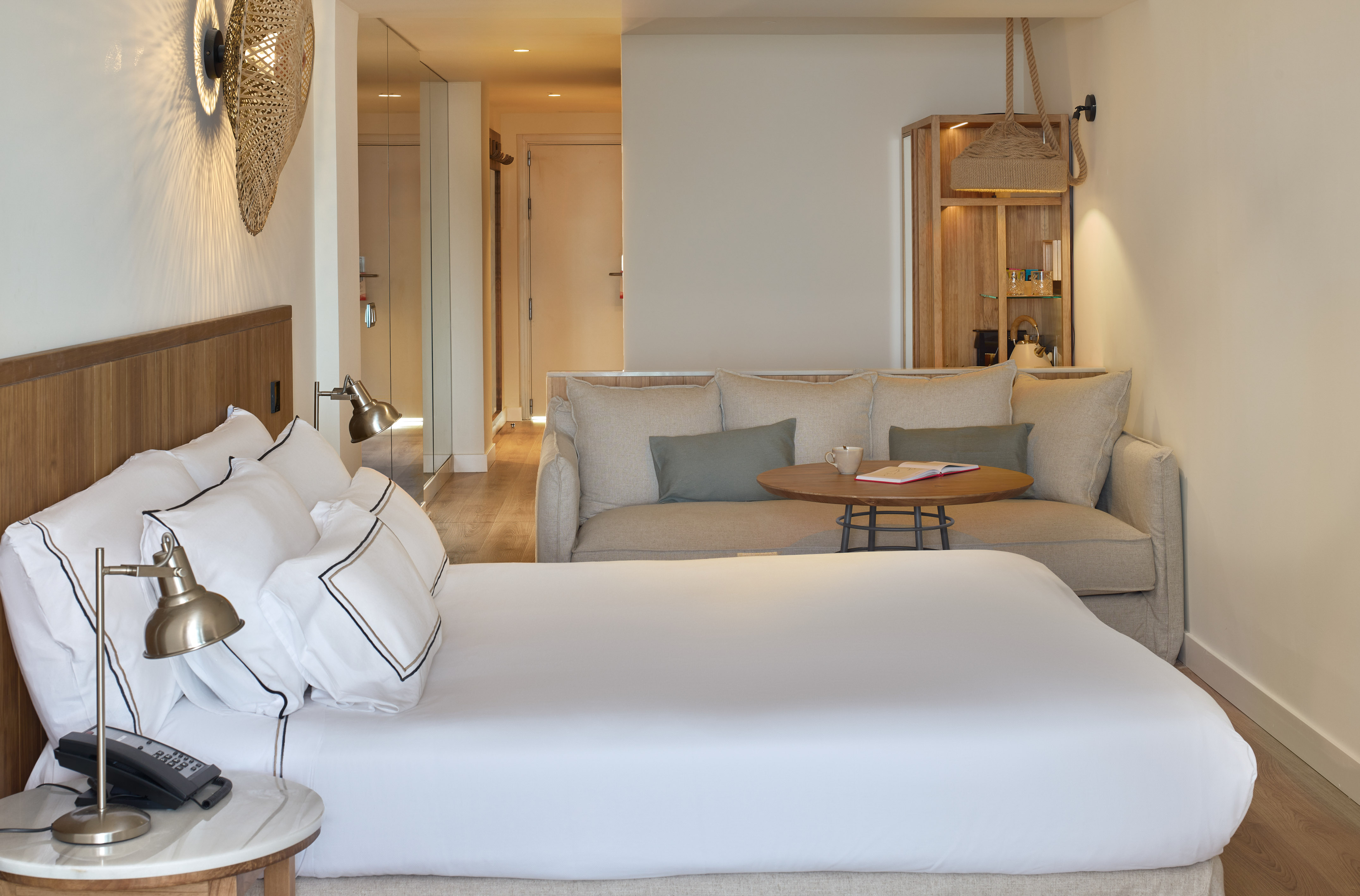 Bright modern hotel room with white bed, sofa, and natural wood furnishings.