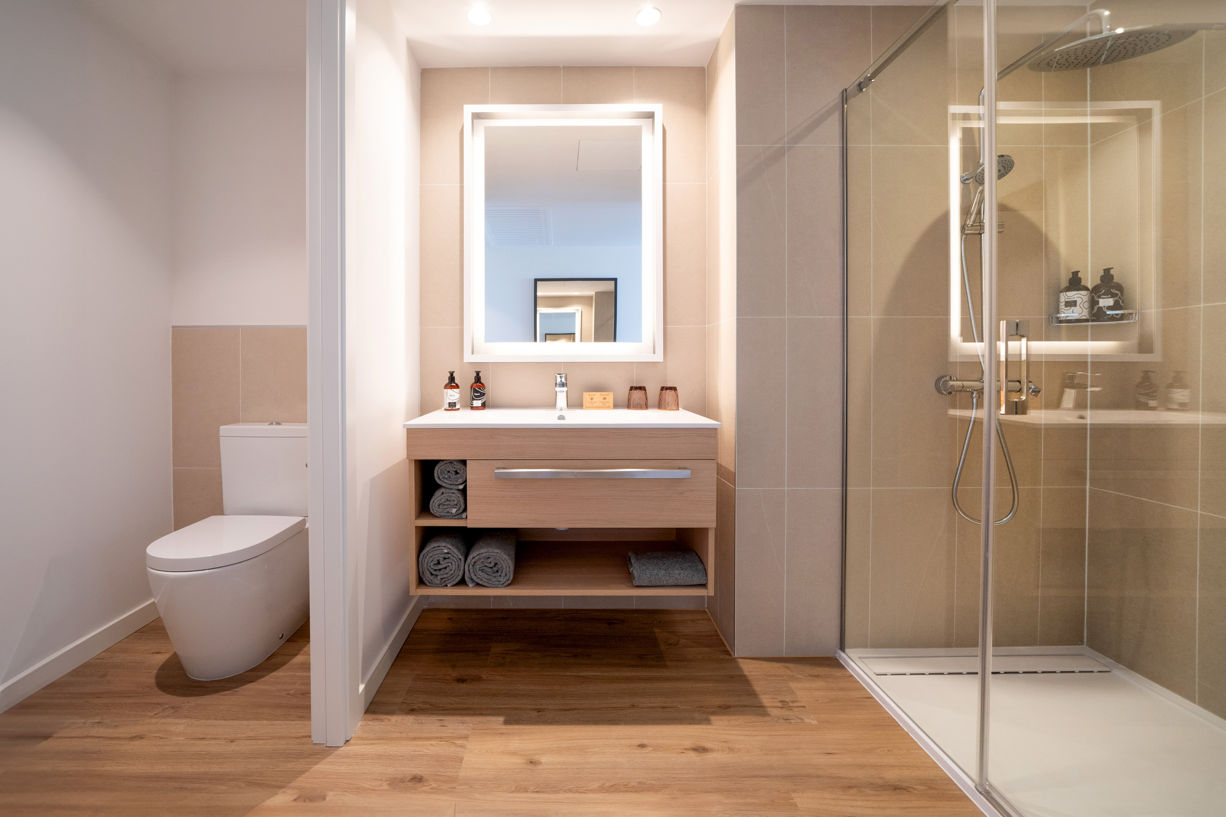 Modern bathroom with wood vanity, backlit mirror, glass shower, and toilet.